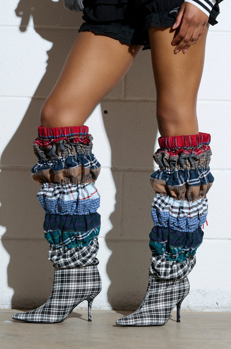 Against a white wall, someone showcases their AZALEA WANG ELODIE MULTI BOXER BOOT with layers of vibrant plaid fabric. The knee-high stiletto heels pair effortlessly with sleek black shorts, creating a bold, fashionable look.