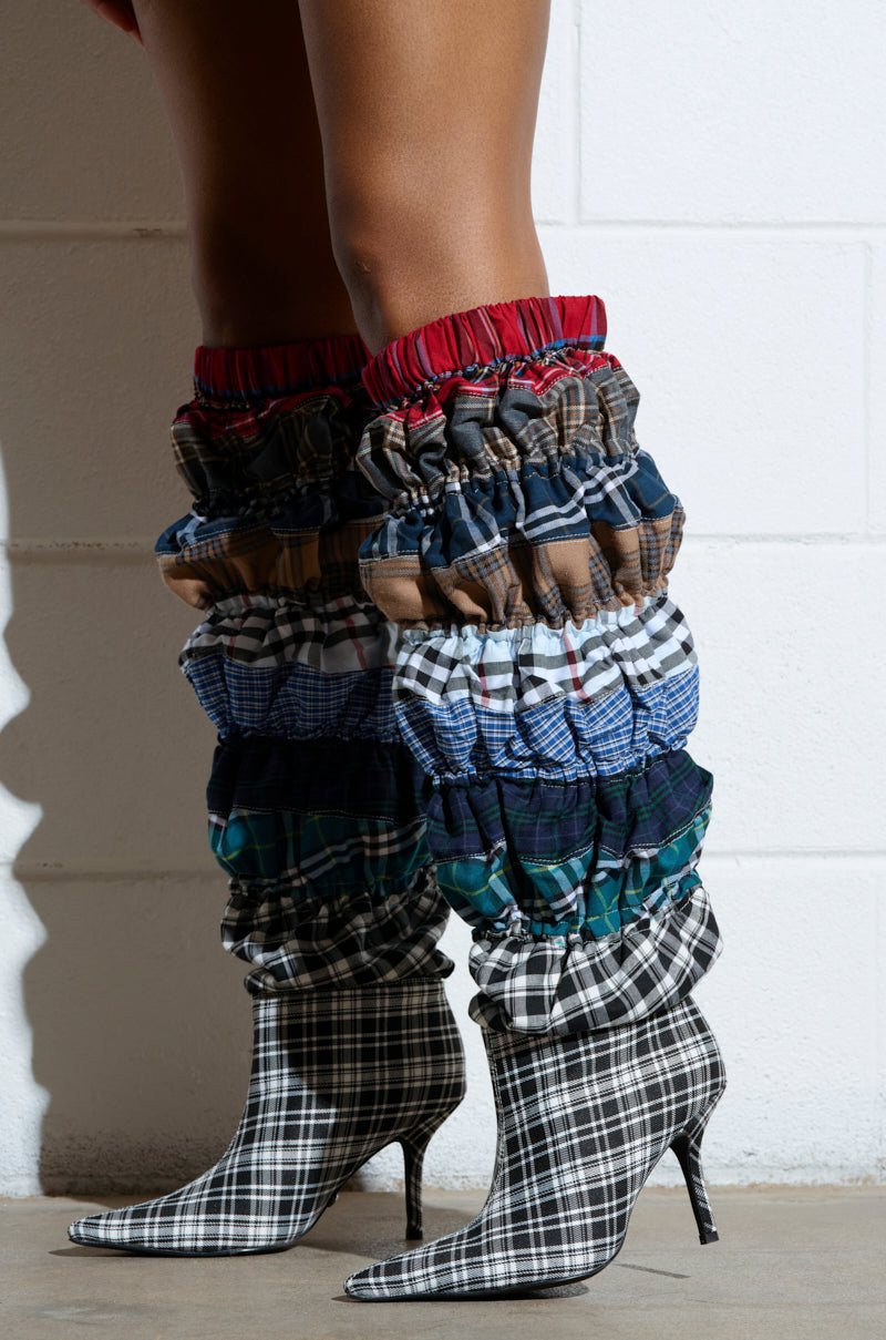 Against a white brick wall, someone wears AZALEA WANG ELODIE MULTI BOXER BOOTS. These slouchy, knee-high boots feature multi-patterned plaid prints in red, brown, blue, and green tones with a pointed stiletto heel that enhances the edgy black and white plaid design.