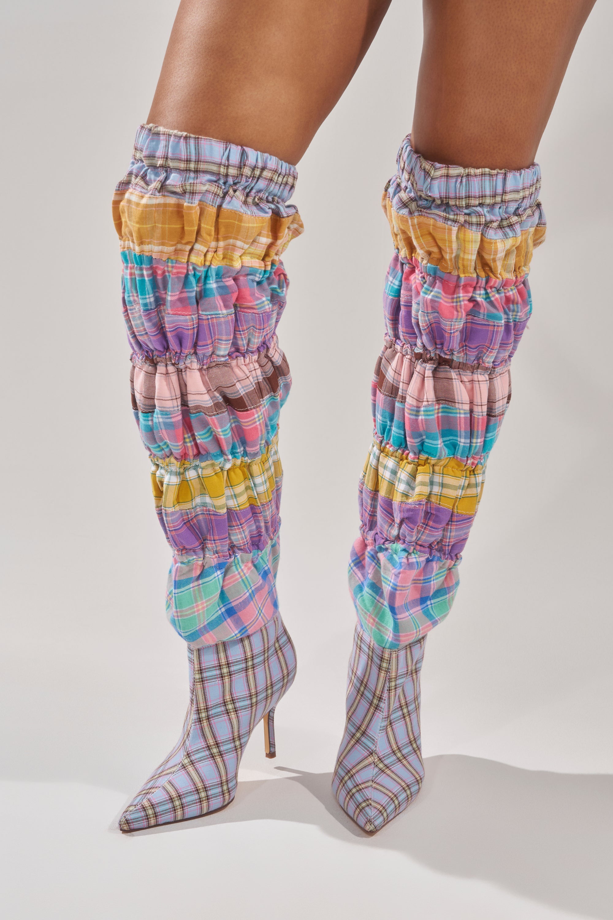 A person wearing the AZALEA WANG ELODIE PINK MULTI BOXER BOOT—thigh-high stiletto boots in pastel pink, purple, yellow, and blue plaid with pointed toes—stands against a plain white background. 0