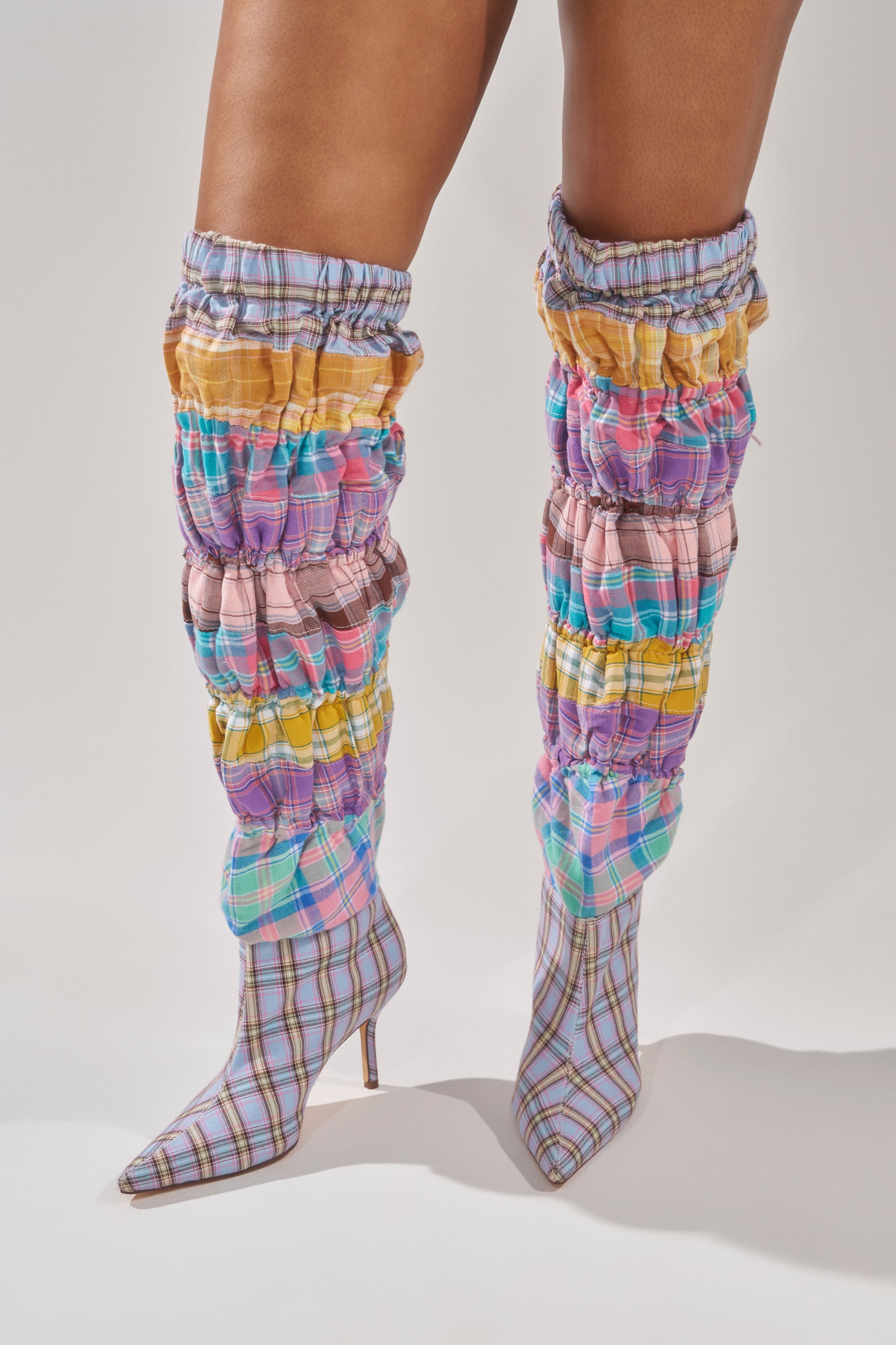 A person stands against a plain background wearing the AZALEA WANG ELODIE PINK MULTI BOXER BOOT, featuring pastel plaid, ruched, knee-high design with pointed toes and stiletto heels. 1
