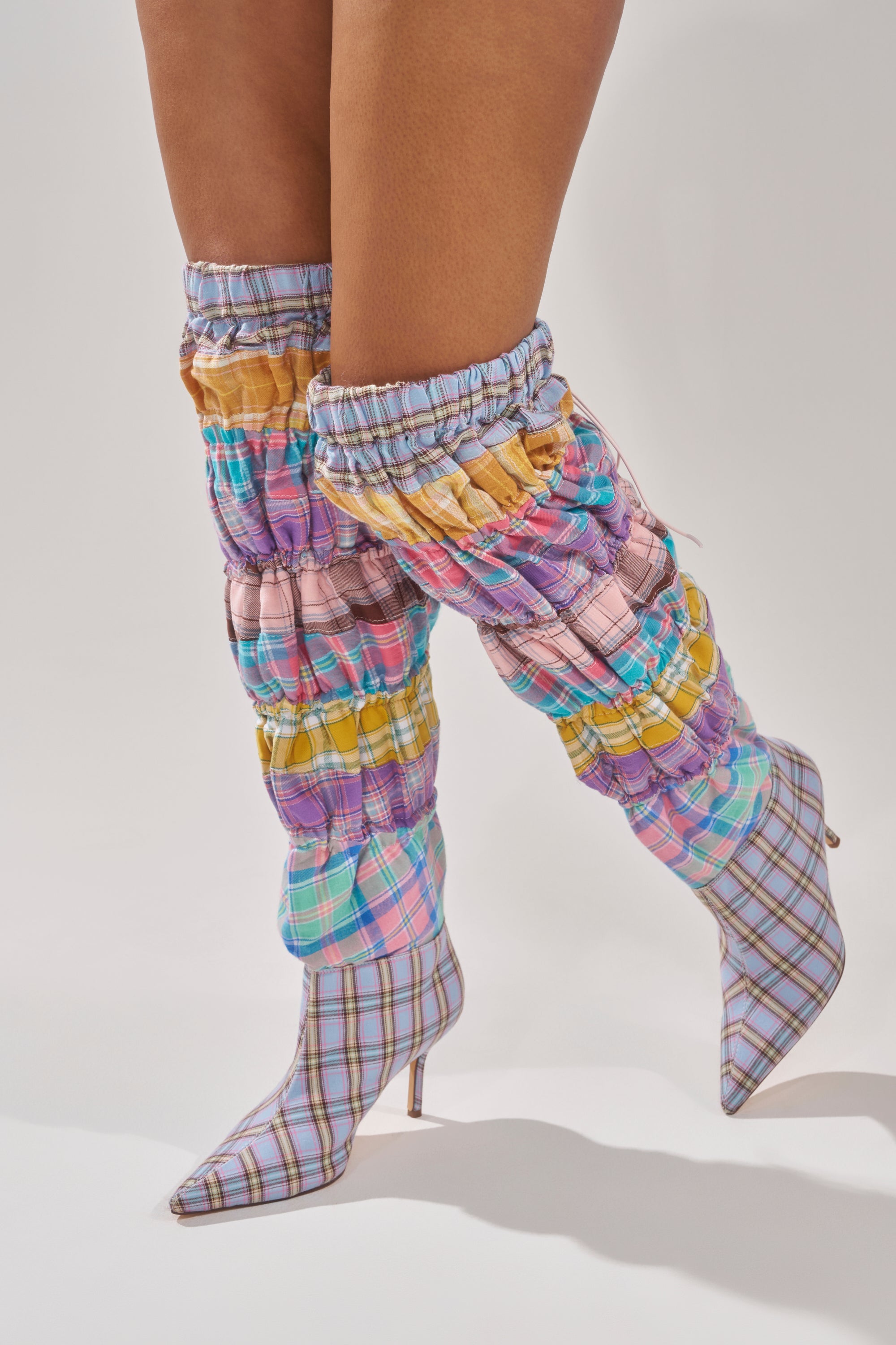 A person wears the AZALEA WANG ELODIE PINK MULTI BOXER BOOT—colorful, ruched, over-the-knee boots with a pastel plaid pattern and pointed toes—while standing against a plain light background. 2