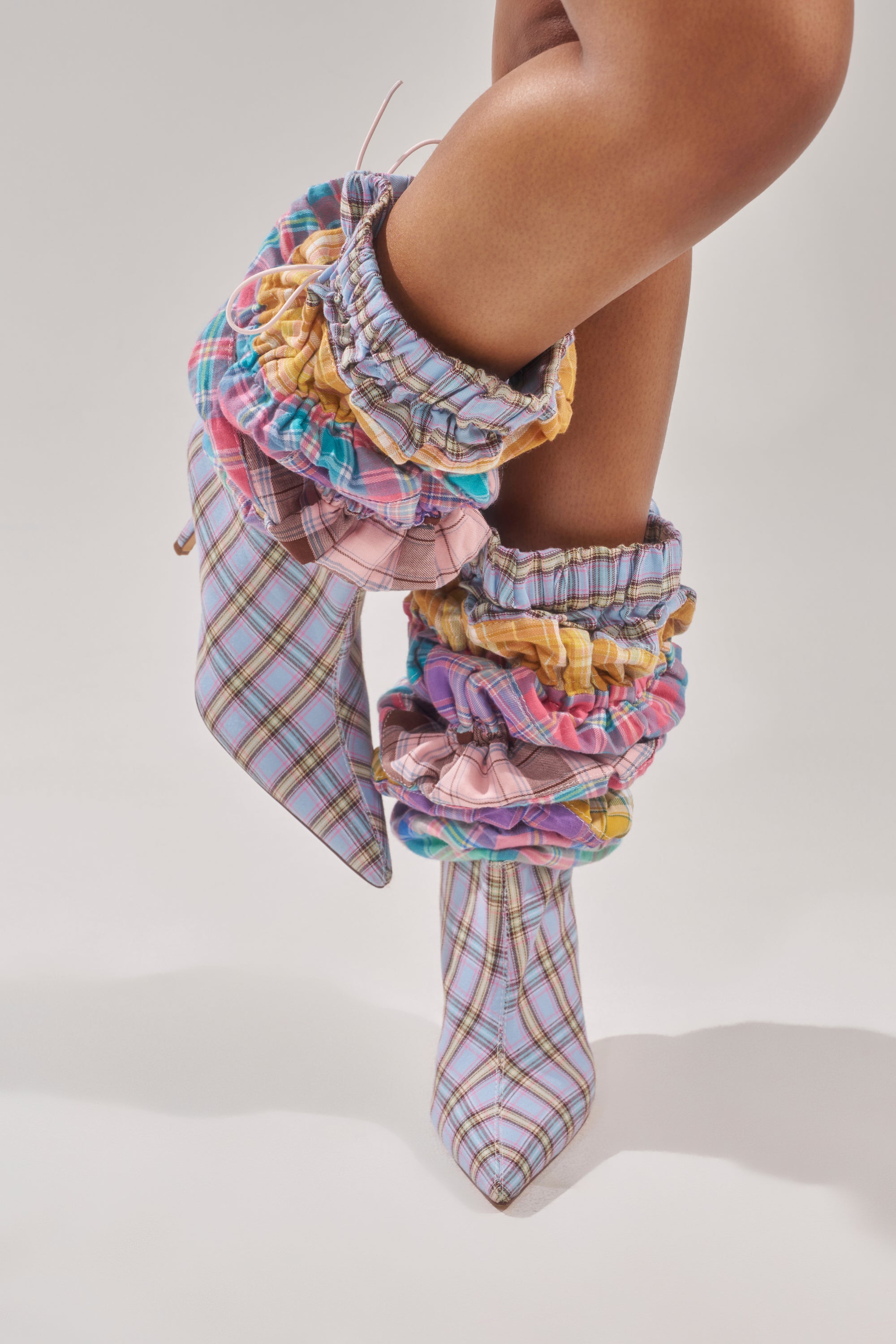 A person stands on tiptoe against a plain light background wearing the AZALEA WANG ELODIE PINK MULTI BOXER BOOT, featuring patchwork pastels and large, colorful, scrunched fabric cuffs around the ankles. 4