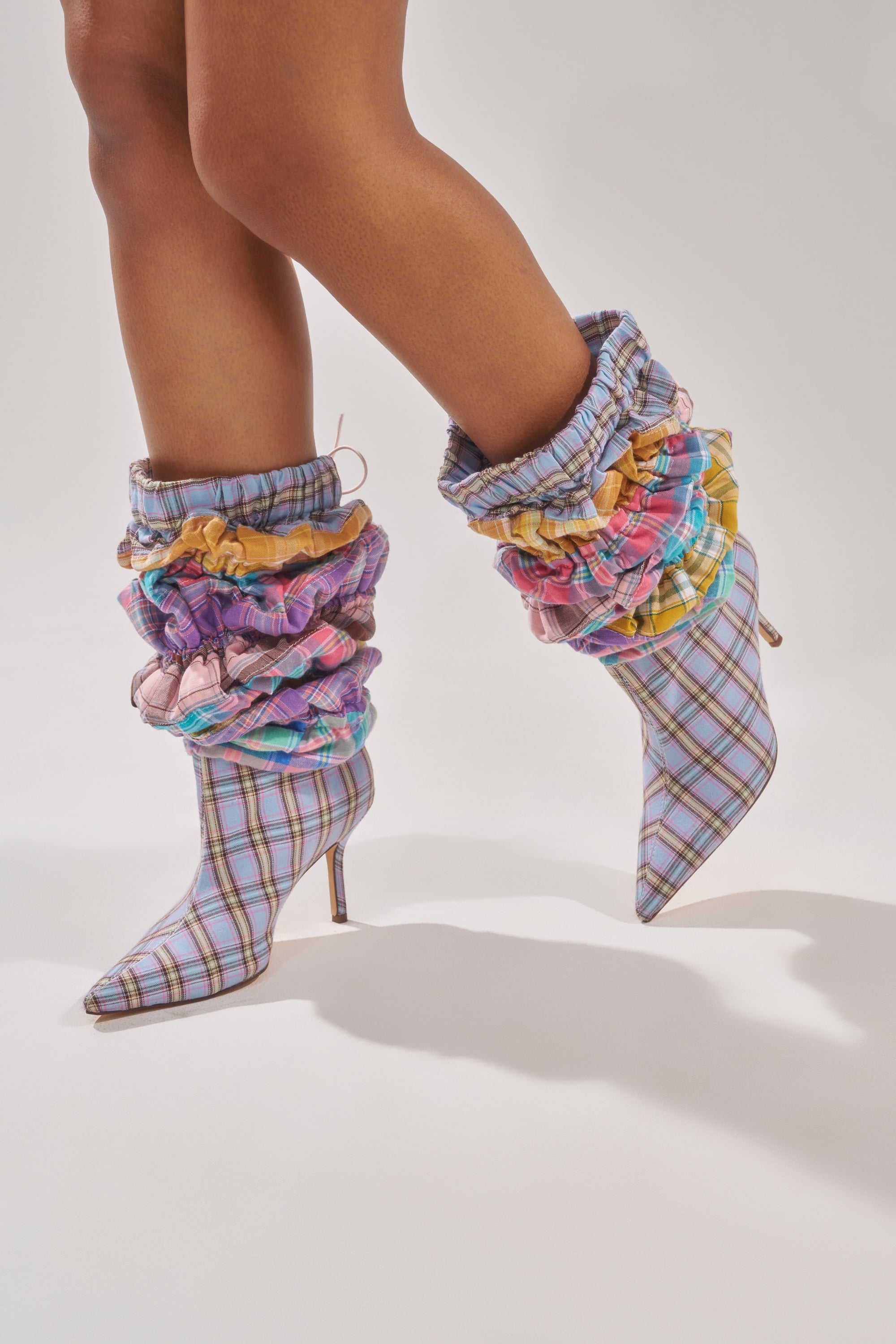 A person wears AZALEA WANG ELODIE PINK MULTI BOXER BOOTS with patchwork pastels, pointed toes, stiletto heels, and colorful ruffled ankle accents. These bold boots stand out against a plain, light background. 5