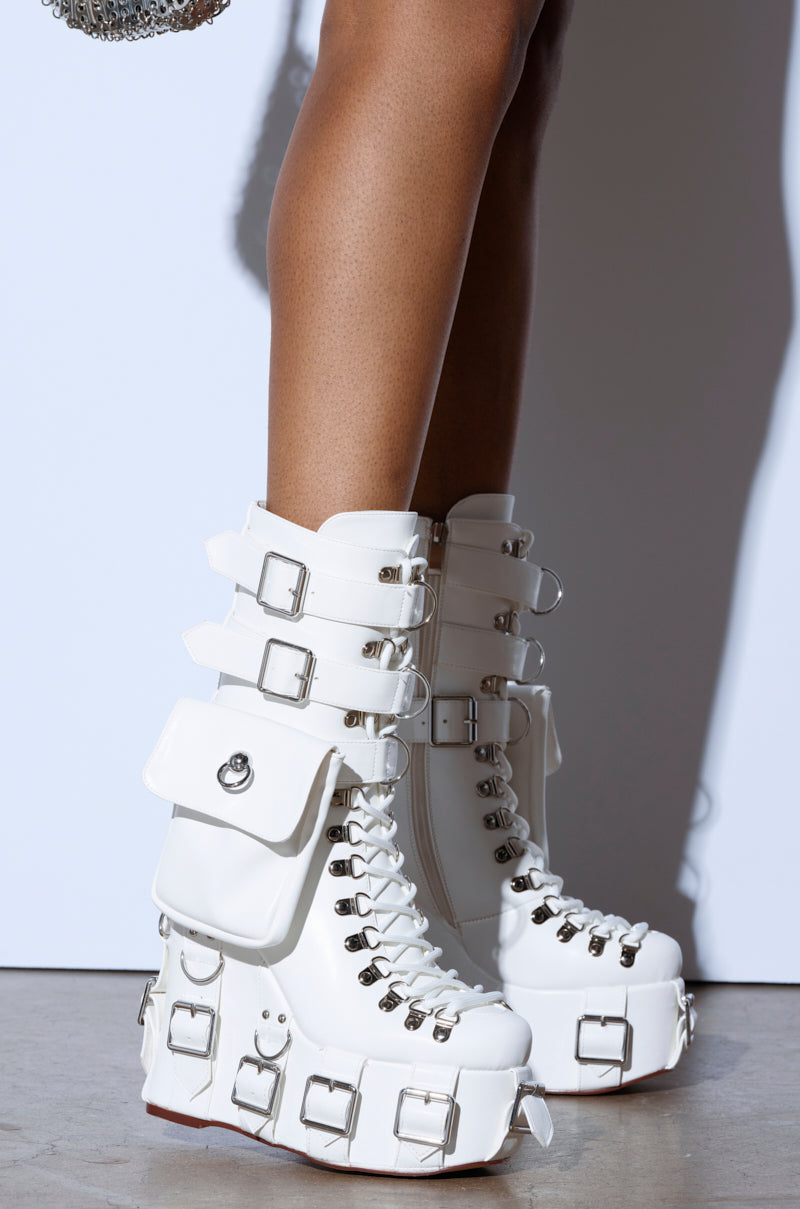 On a beige surface, someone wears AZALEA WANG ELOWYN WHITE EDGY WEDGE BOOT—white knee-high platform boots made from faux leather, featuring decorative buckles, numerous laces, metal details, and a small pouch. A plain white wall in the background complements the striking boots.