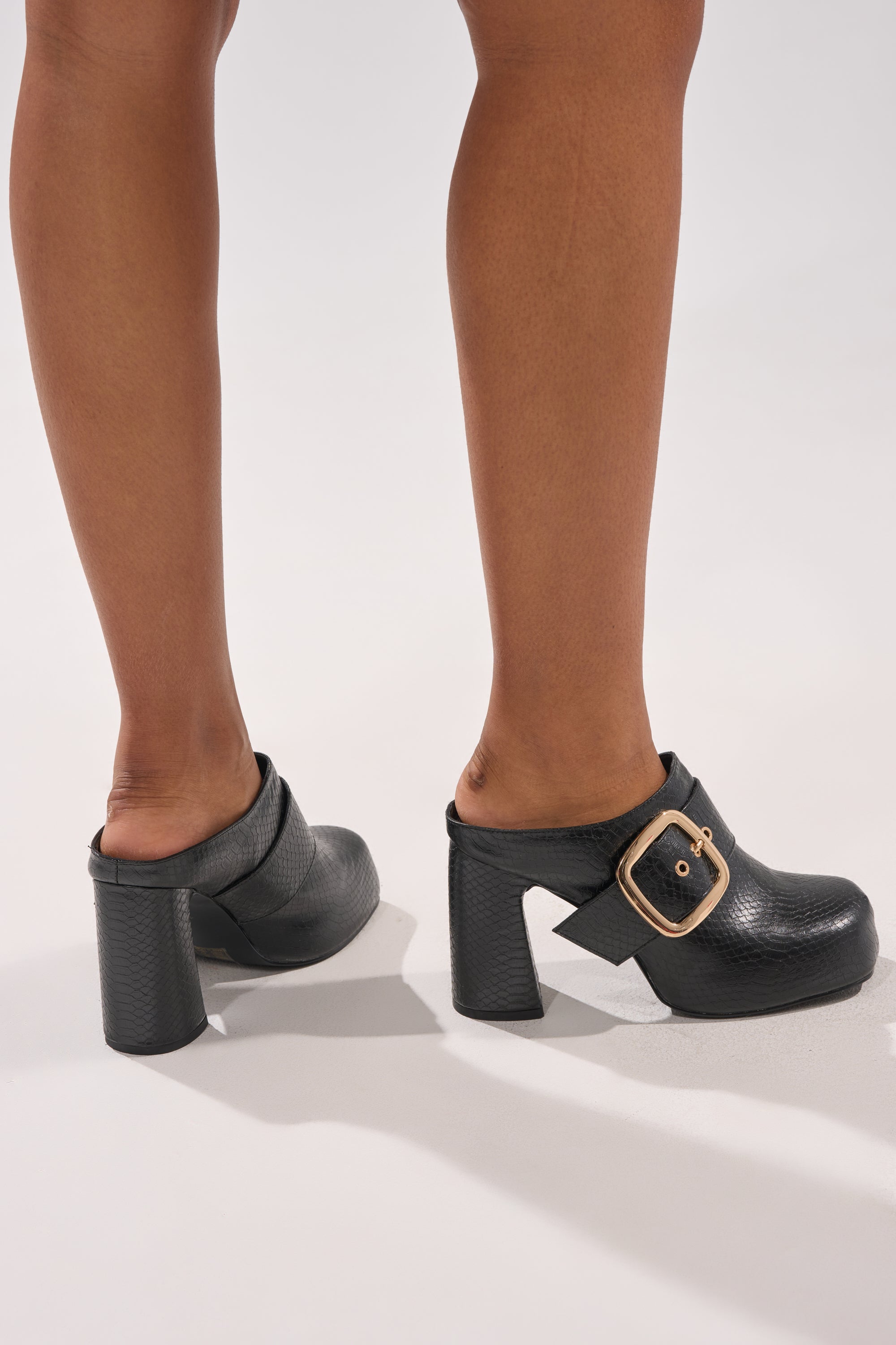 A person wearing the AZALEA WANG ELYSIA BLACK PU ROUND TOE MULE with crocodile print and large gold buckles stands on a light gray surface, showing only their lower legs and feet. 2