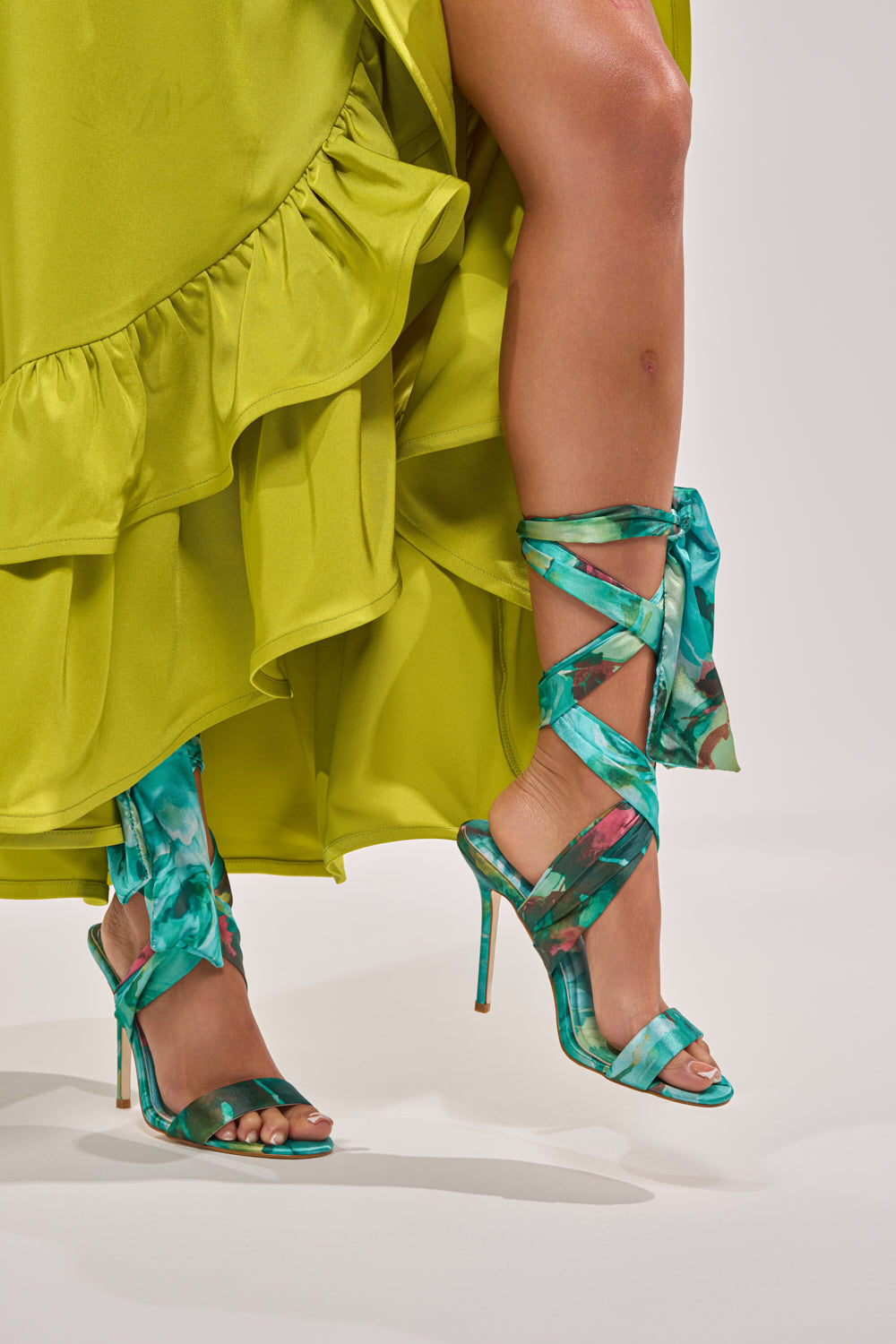 A person in a bright green ruffled dress stands with one leg forward, showcasing the AZALEA WANG ELZON GREEN SATIN STRAPPY SANDAL with teal floral details, wrap-around ankle straps, and a sleek stiletto heel. 0