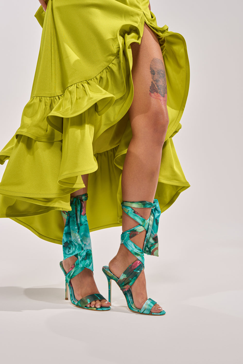 A person wears a bright yellow ruffled dress and the AZALEA WANG ELZON GREEN SATIN STRAPPY SANDAL. They have a tattoo on their thigh. The background is plain and light-colored. 2