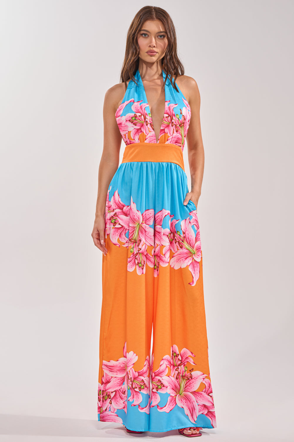 A woman wears the ON VACA TIME HALTER FLORAL PRINT JUMPSUIT, a sleeveless halter-neck jumpsuit with a deep V-neck, bright blue and orange color block design, and bold pink floral prints at the waist and on the pant legs. She stands facing forward. 0
