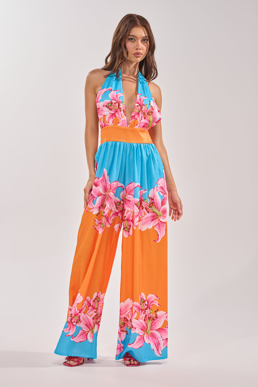 Woman in the ON VACA TIME HALTER FLORAL PRINT JUMPSUIT featuring a deep neckline, bold orange and blue panels, and large pink floral prints, posed against a plain light background. 1