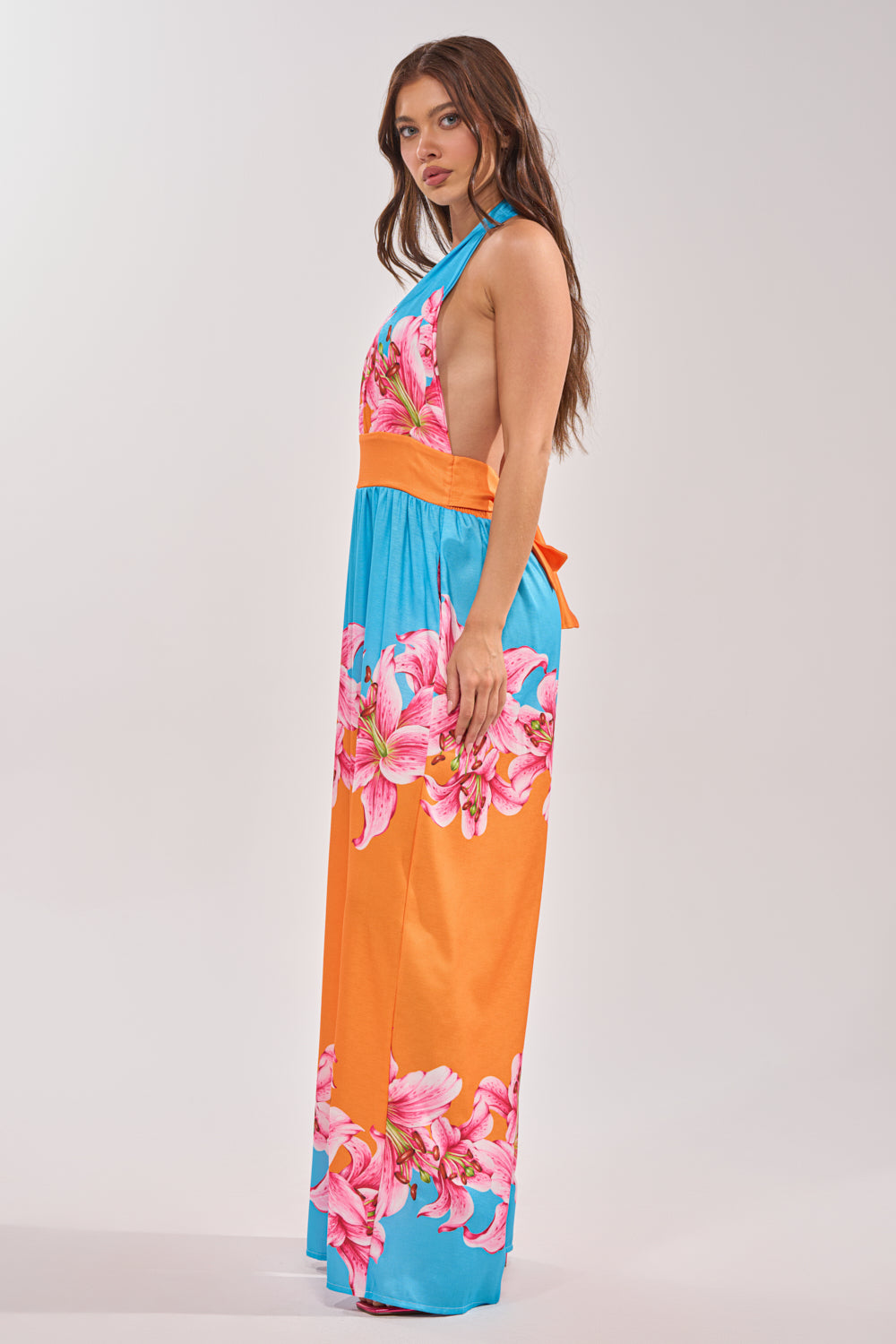 A woman stands sideways, looking at the camera, wearing the ON VACA TIME HALTER FLORAL PRINT JUMPSUIT with a colorful floral pattern, low back, and flowy silhouette against a plain light-colored background. 2