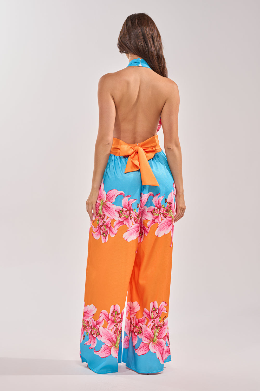 A woman faces away, wearing the ON VACA TIME HALTER FLORAL PRINT JUMPSUIT. This vibrant, backless halter has wide legs, pink floral prints in orange and blue hues, and a large waist bow for a bold tropical style. 3