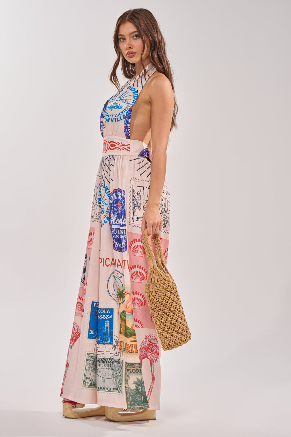A woman with long brown hair stands sideways against a plain background, wearing the CATCH YOU LATER HALTER PRINTED JUMPSUIT with vibrant stamp and postcard prints, platform sandals, and carrying a woven tan bag. 2