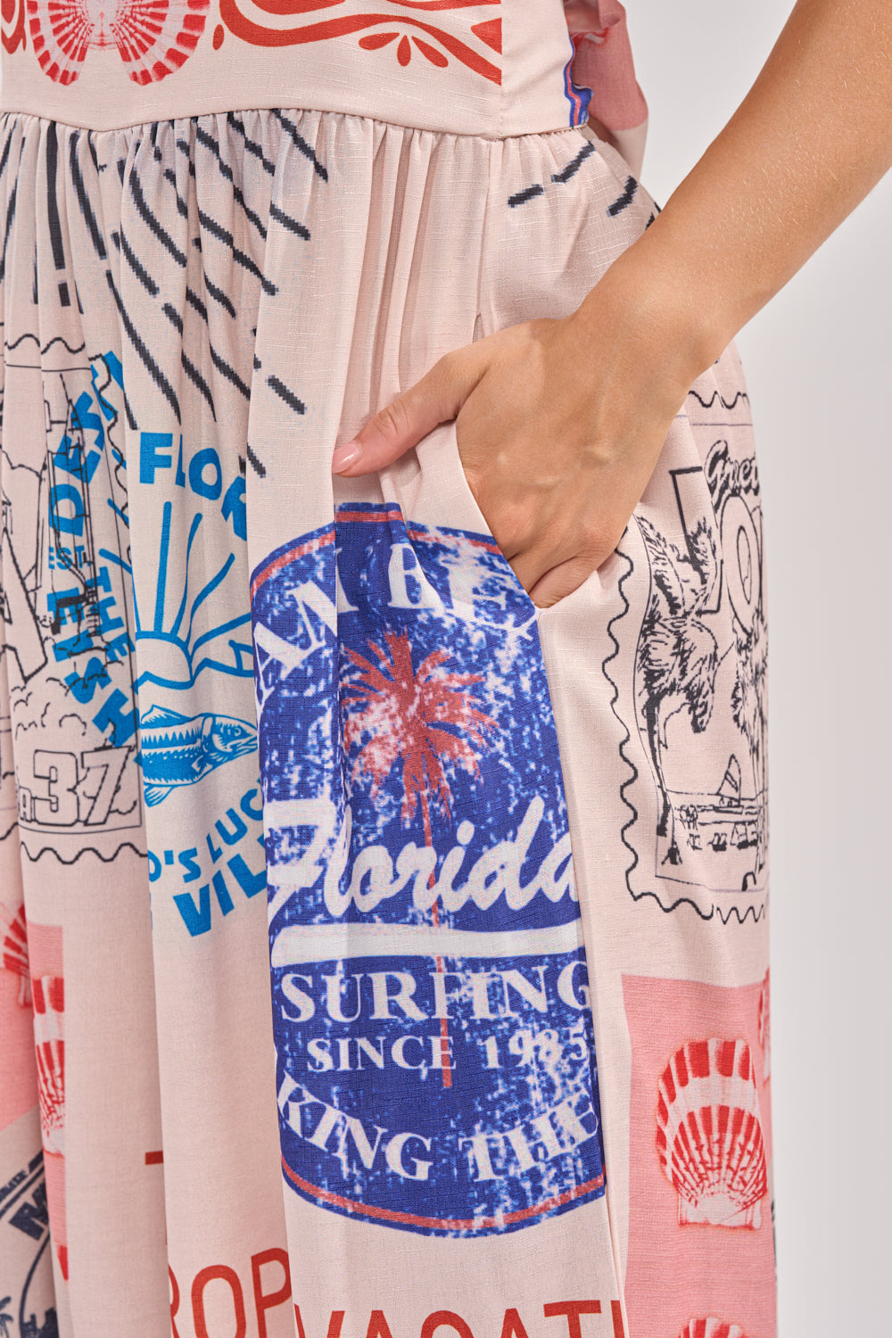 A hand rests in the pocket of the CATCH YOU LATER HALTER PRINTED JUMPSUIT, a boho-chic piece with light fabric covered in colorful, vintage-style travel stamps and postcard graphics, including one labeled "Florida. 6