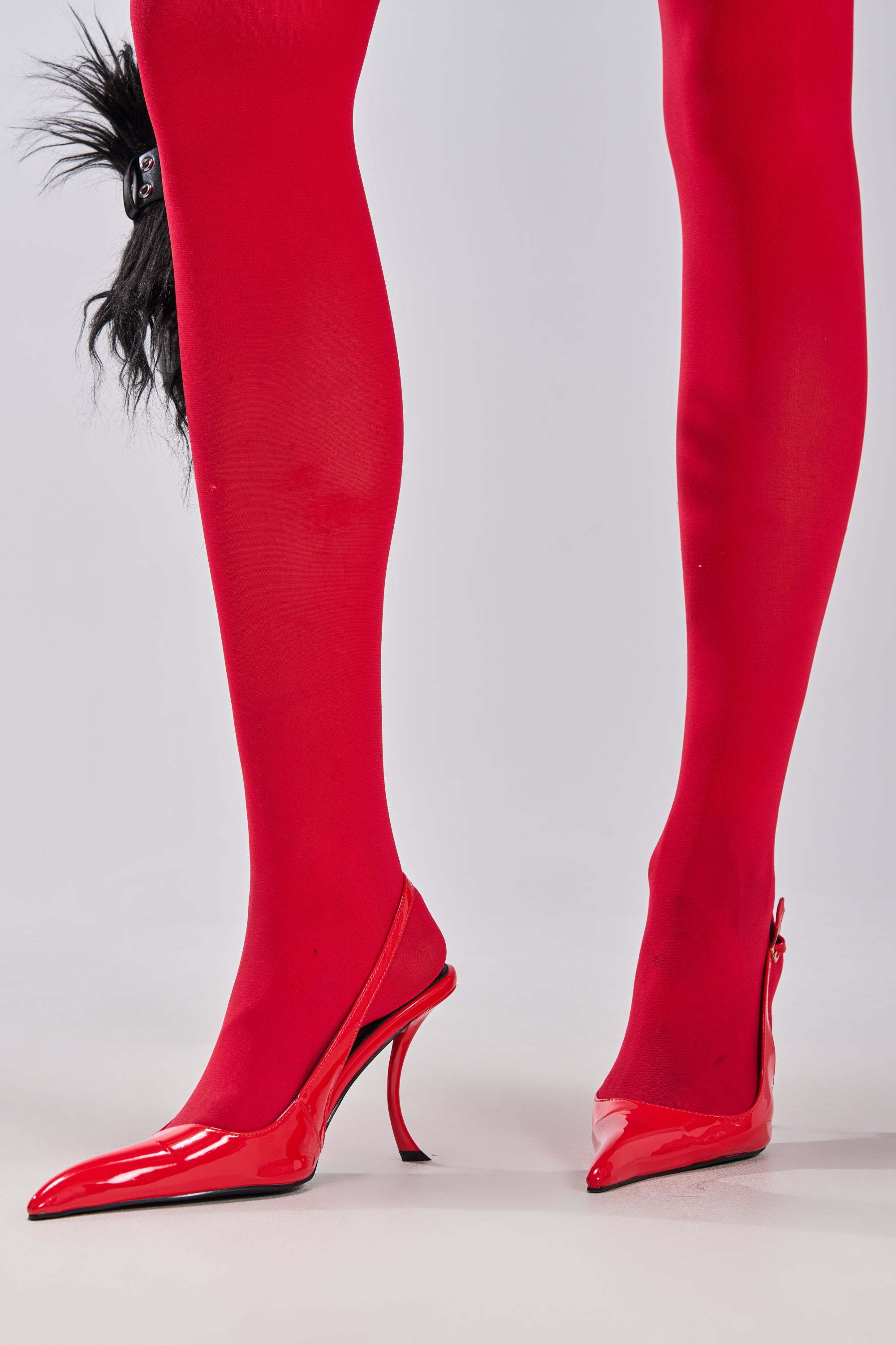 Wearing bright red tights and the AZALEA WANG EMANUELA RED SHARP TOE SLING BACK PUMP with a sleek stiletto heel, a person stands on a light gray floor. A black furry accessory is partially visible on the left. 3