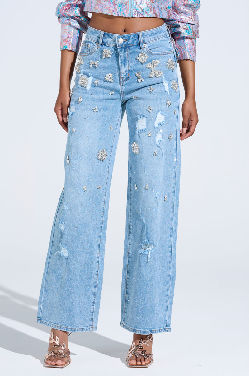 A person wears IN YOUR DREAMS RHINESTONE DENIM JEANS featuring silver floral patterns and distressed details. They pair them with a colorful patterned cropped jacket and open-toe sandals against a plain white canvas.