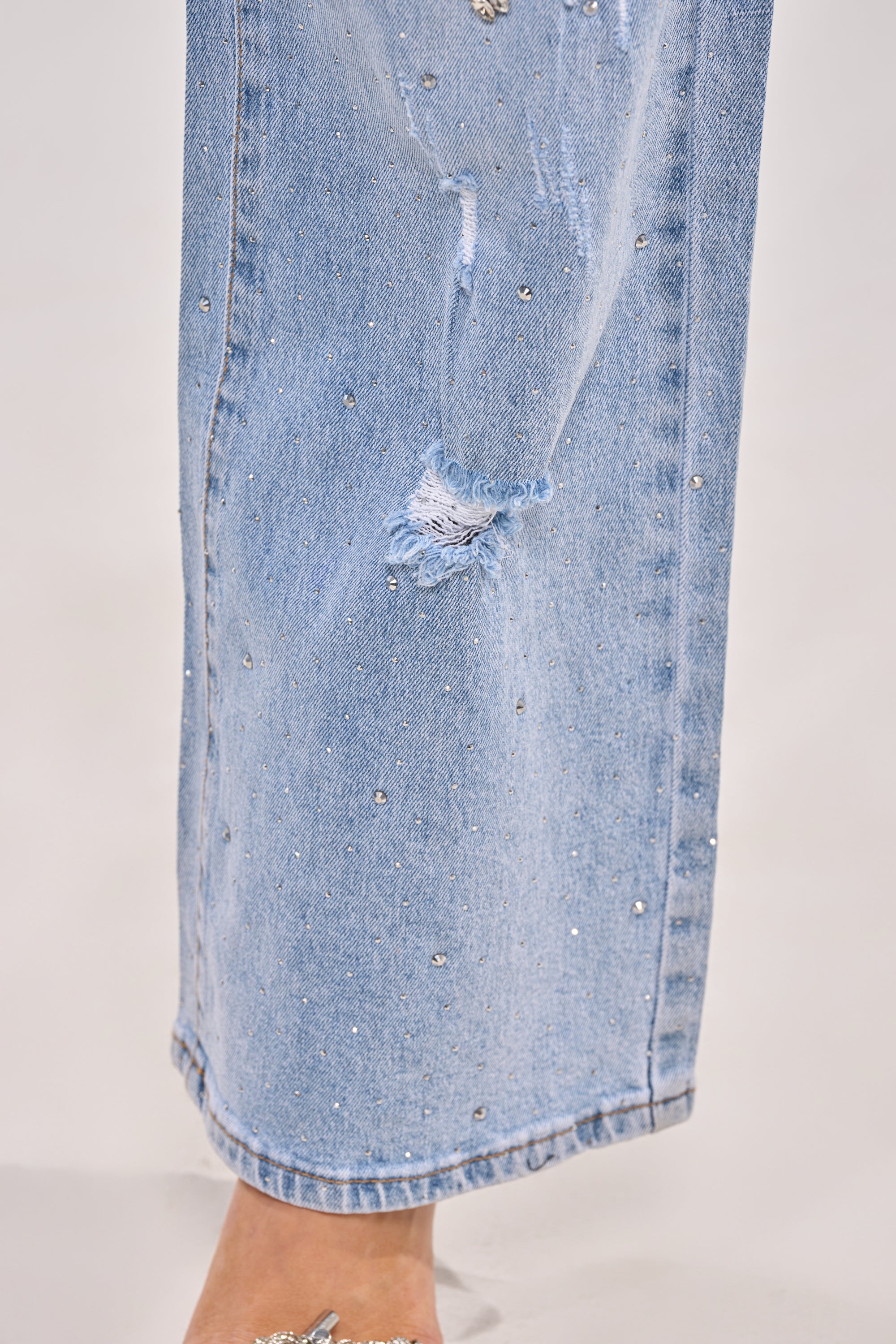 A close-up of the IN YOUR DREAMS RHINESTONE DENIM jeans shows small light blue rhinestones and a subtle knee rip. These bedazzled jeans feature a relaxed, straight-leg fit for effortless style. 9