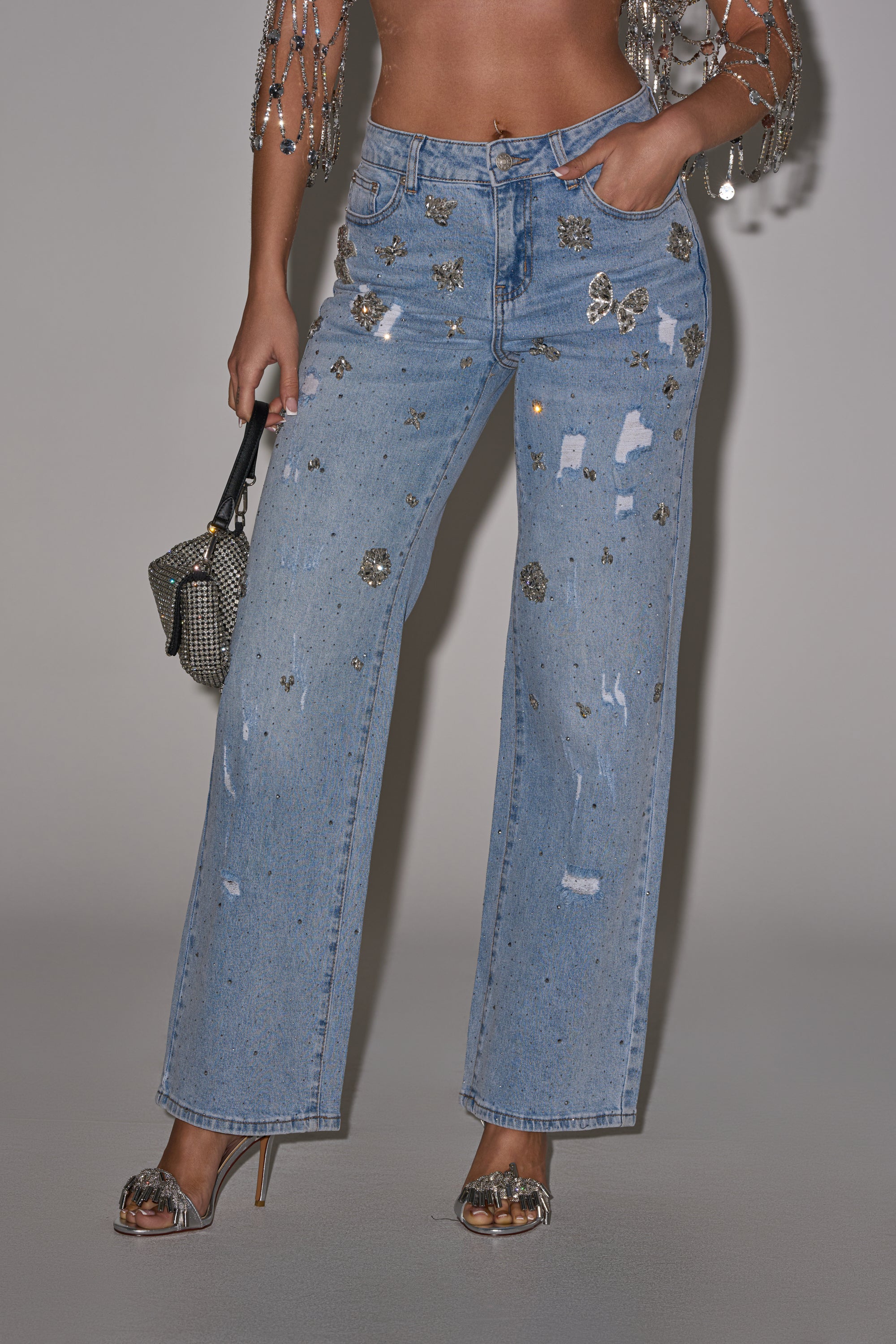 A person wears the IN YOUR DREAMS RHINESTONE DENIM—high-waisted, light blue wide-leg jeans with rhinestones and star patches—paired with silver heels, a glittery mini bag, and a beaded top draped over their shirtless torso. 0