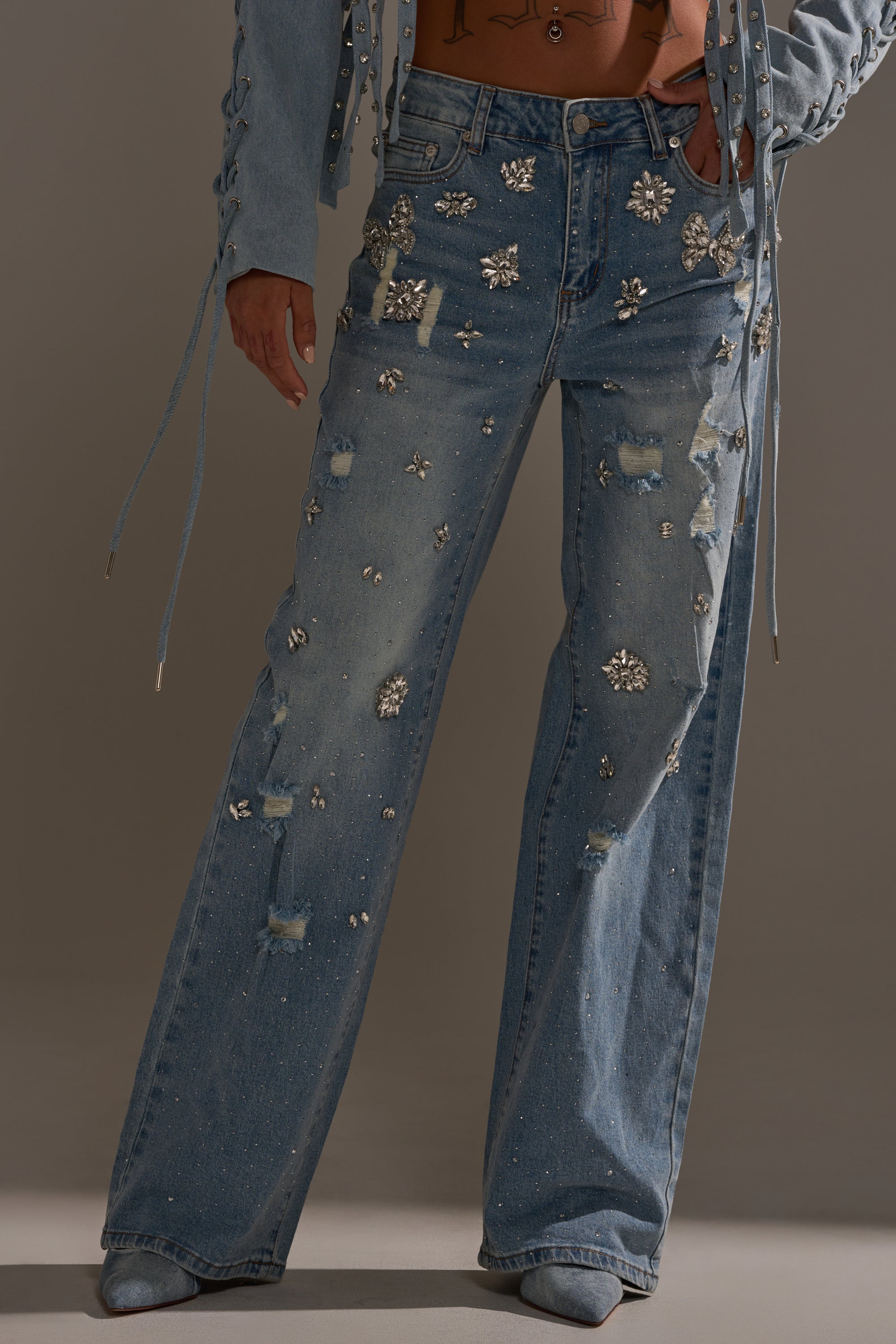 Wearing IN YOUR DREAMS RHINESTONE DENIM JEANS with gem-like embellishments and a matching light blue jacket with long, dangling strings, this person sports trendy wide-leg denim. Their face is not visible.