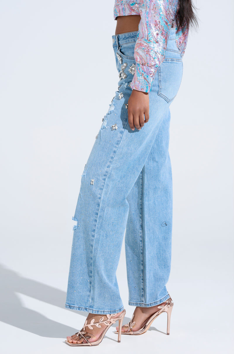 Someone wearing IN YOUR DREAMS RHINESTONE DENIM JEANS and a vibrant long-sleeve top stands in profile. They pair it with strappy high heels featuring a metallic leaf design, adding sparkle against the light gray backdrop.