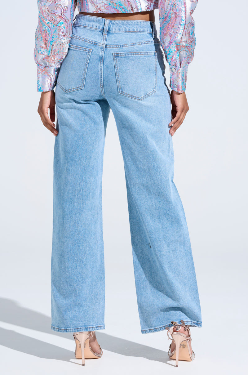 A person wearing the IN YOUR DREAMS RHINESTONE DENIM JEANS in a light blue, wide-leg silhouette and a patterned blouse stands with their back to the camera, styled with nude heels featuring ankle ties. The plain, light-colored background subtly complements this elegant ensemble.