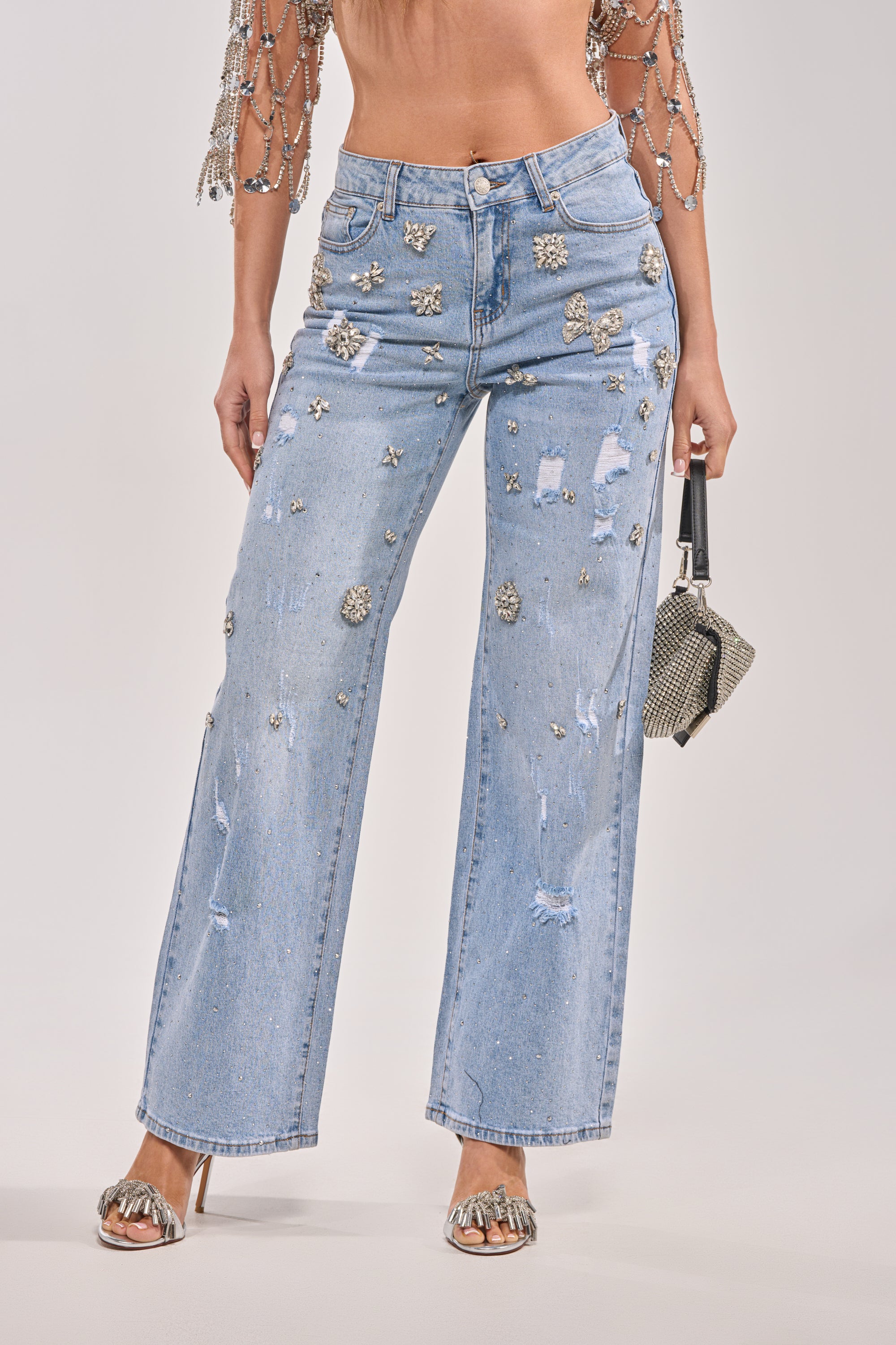 A woman wears the IN YOUR DREAMS RHINESTONE DENIM jeans in light blue with a cropped beaded top, silver heeled sandals, and a small metallic handbag. 3