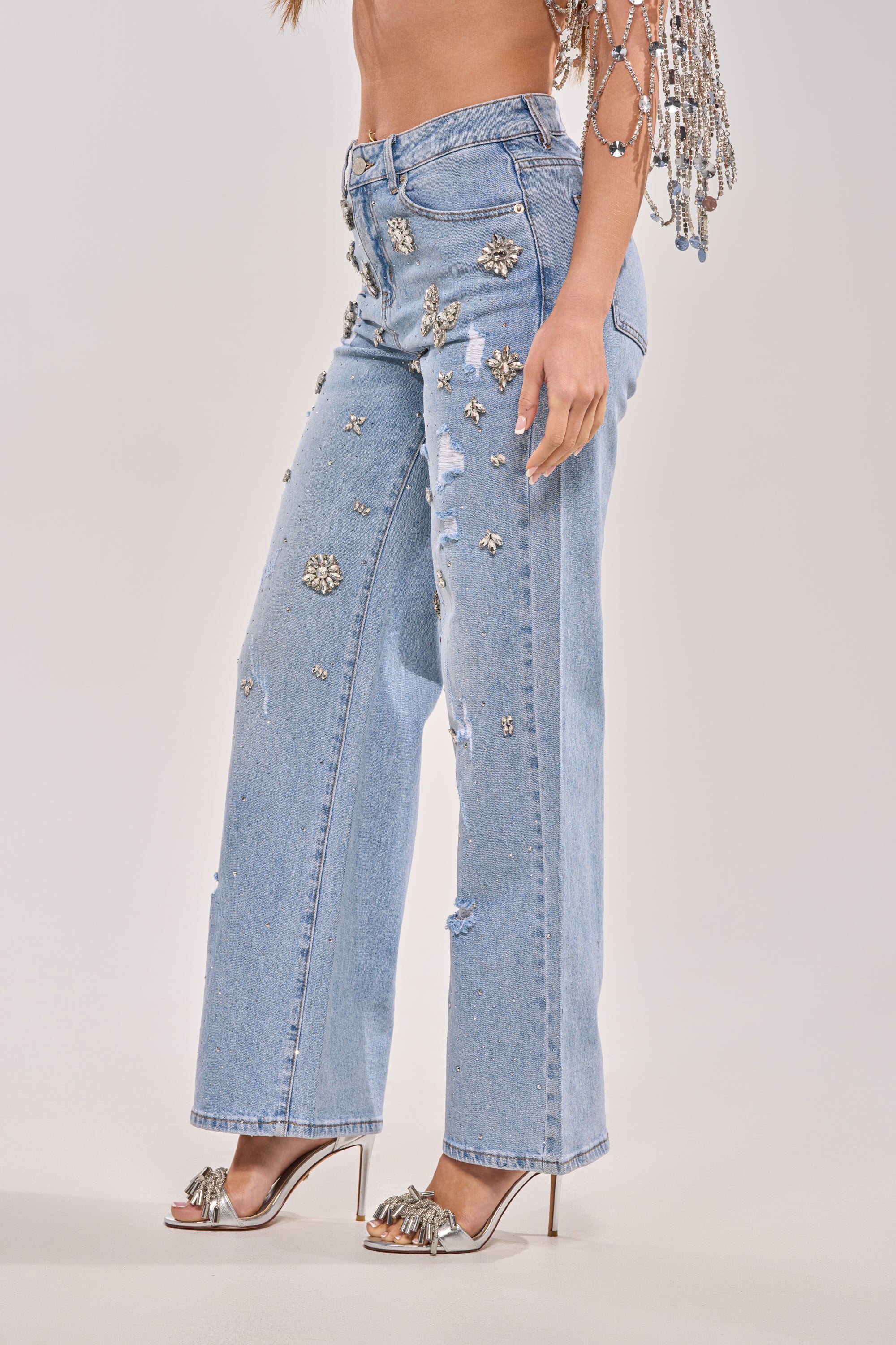 A person wears the IN YOUR DREAMS RHINESTONE DENIM wide-leg jeans in light blue with bedazzled details, paired with silver high-heeled sandals and a beaded top. The image is cropped at the shoulders. 4