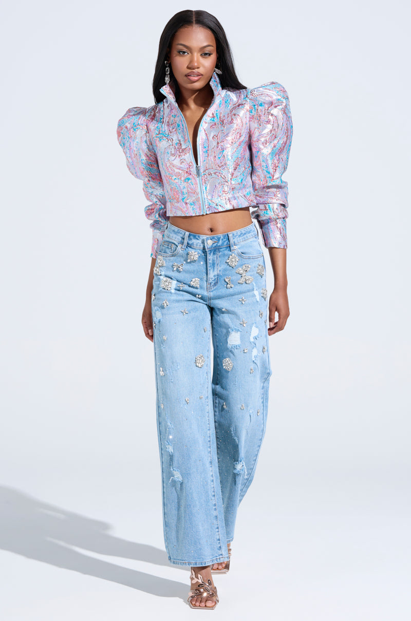 A woman in a colorful, patterned jacket and IN YOUR DREAMS RHINESTONE DENIM JEANS with wide legs and floral embellishments stands against a plain light background, wearing silver heels.