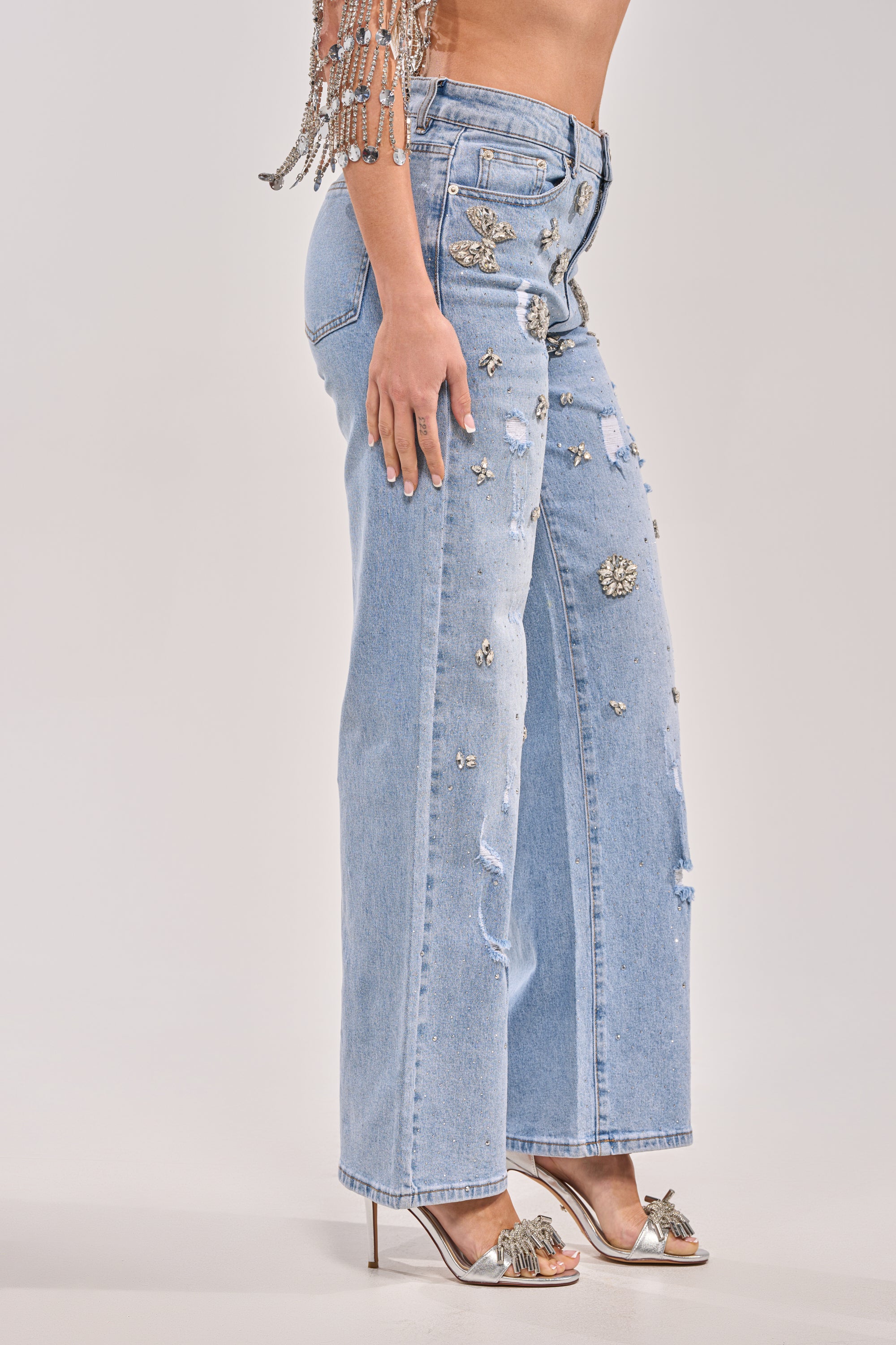 Someone models the IN YOUR DREAMS RHINESTONE DENIM featuring silver embellishments, styled with silver high-heeled sandals and a beaded, sparkly top against a plain light background. 5