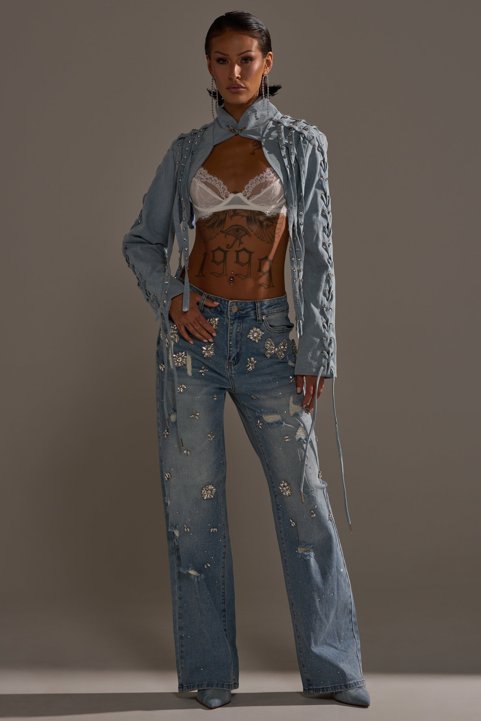 A woman with short dark hair and visible torso tattoos poses in a studio wearing the IN YOUR DREAMS RHINESTONE DENIM JEANS, paired with a light denim jacket featuring braided details and a white lace bralette.