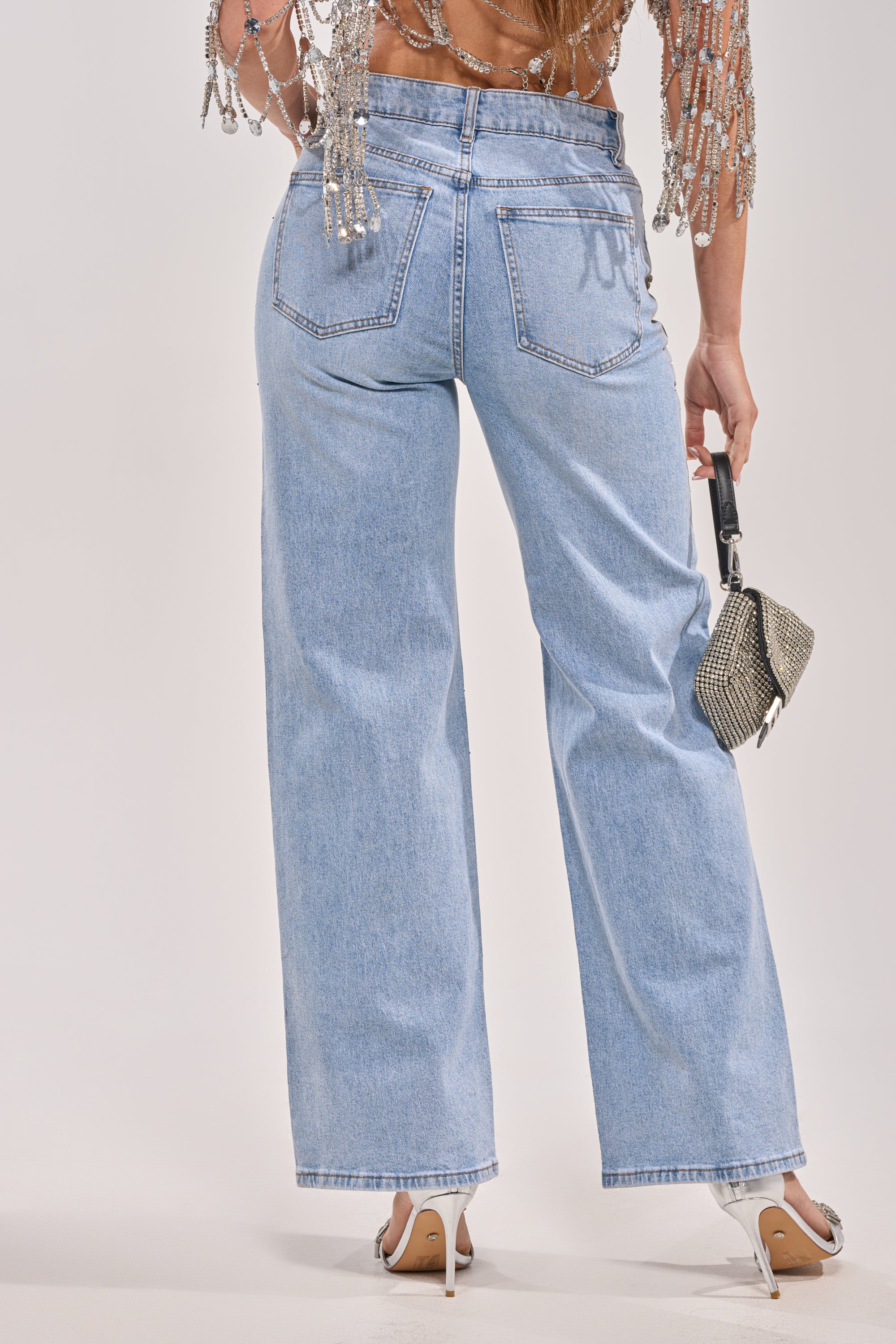 A person is shown from the back in IN YOUR DREAMS RHINESTONE DENIM, paired with a beaded top and high heels, holding a small silver handbag by their side against a plain background. 6