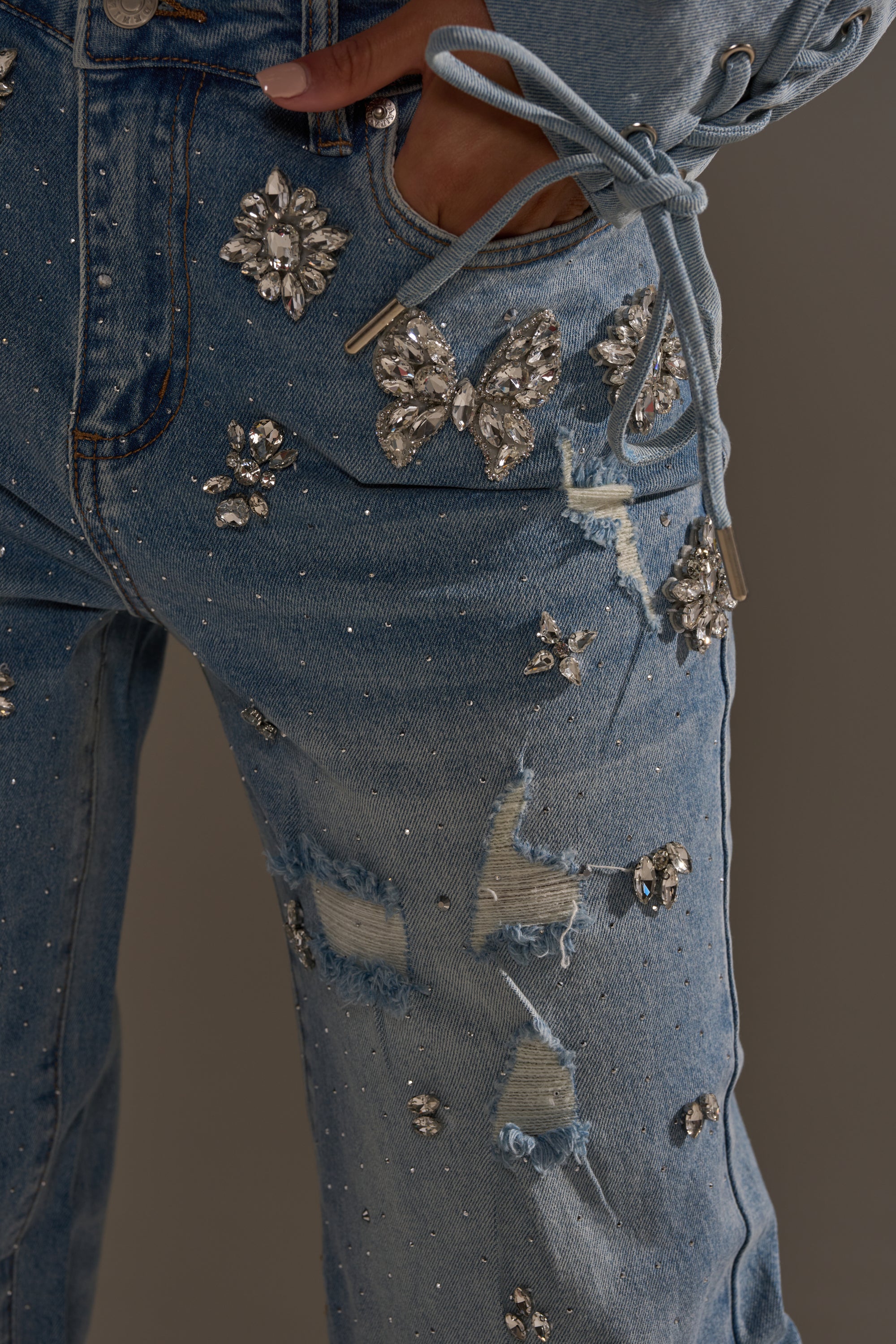 Close-up of the IN YOUR DREAMS RHINESTONE DENIM JEANS featuring butterfly and flower rhinestone accents, distressed patches, and a hand in the front pocket. A matching top's drawstring is visible at the waist, showcasing these trendy embellished jeans.