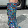 Wearing the AZALEA WANG EMBER RHINESTONE MULTI STILETTO BOOT, a person stands confidently on a crosswalk, knee-high blue boots adorned with sparkling gems that catch the light, while a vehicle is visible in the background.