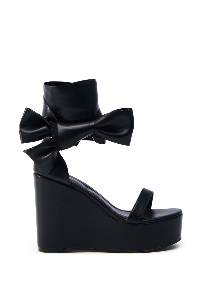 Side view of the AZALEA WANG EMEKA BLACK WEDGE SANDAL showcasing a sleek faux leather design with a black platform sole, thick ankle strap featuring a large bow, comfort cushion insole, and elegant toe strap.