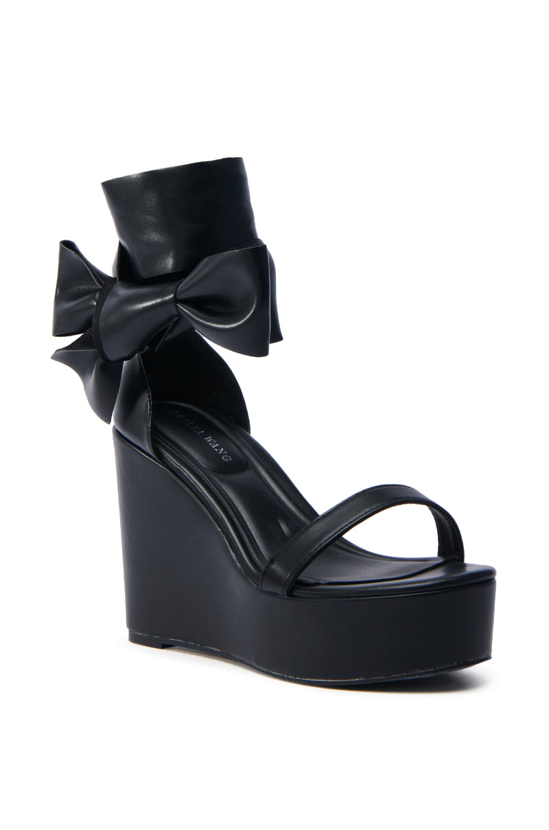 The AZALEA WANG EMEKA BLACK WEDGE SANDAL features a stylish high platform faux leather design with an ankle strap, beautifully accented by a large black bow on the side, making it perfect for any fashion-forward individual.