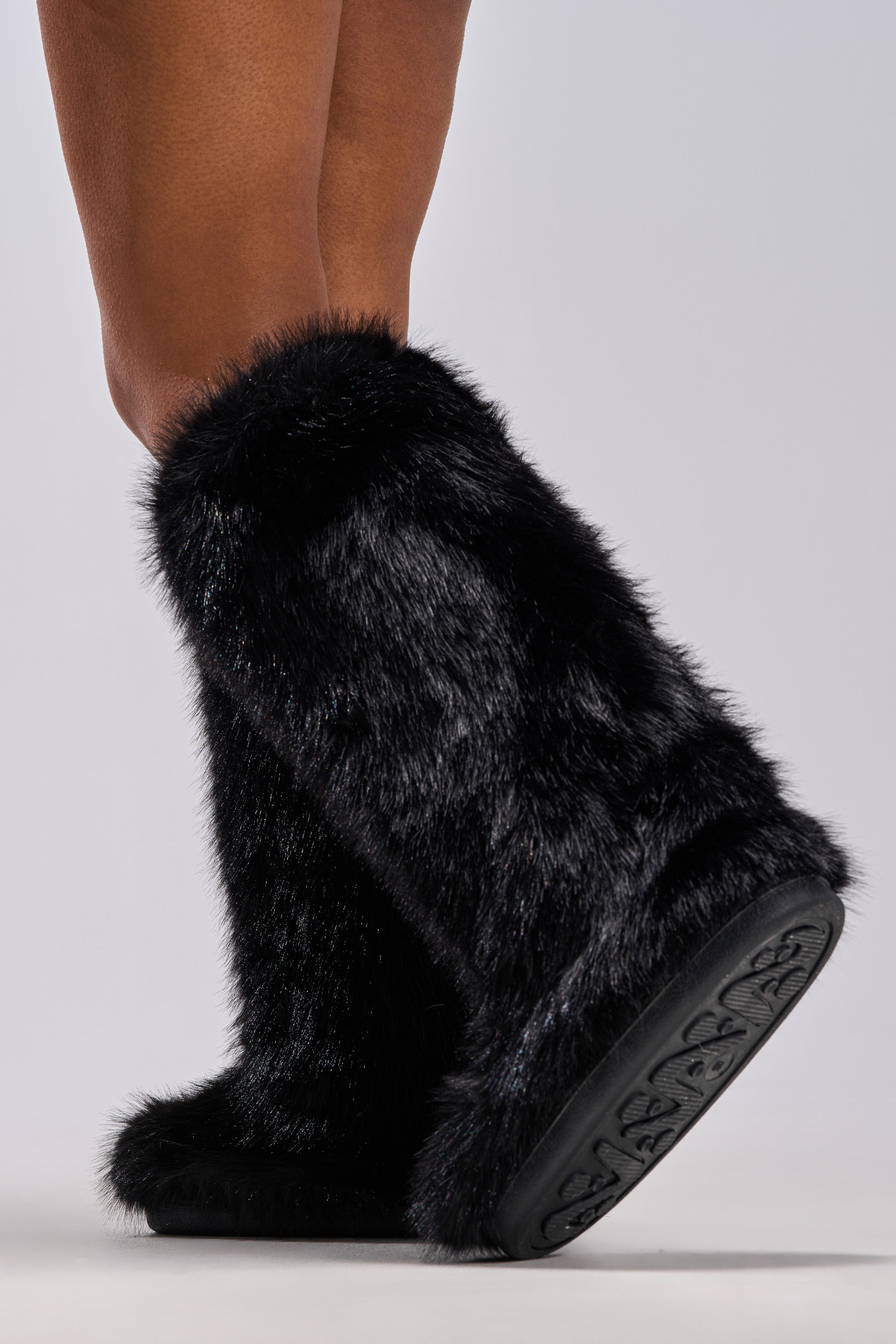 A person wears the AZALEA WANG EMIR BLACK FLAT FUR KNEE HIGH BOOT, featuring thick faux fur and patterned rubber soles, standing on a light grey background. Only their lower legs and feet are visible.