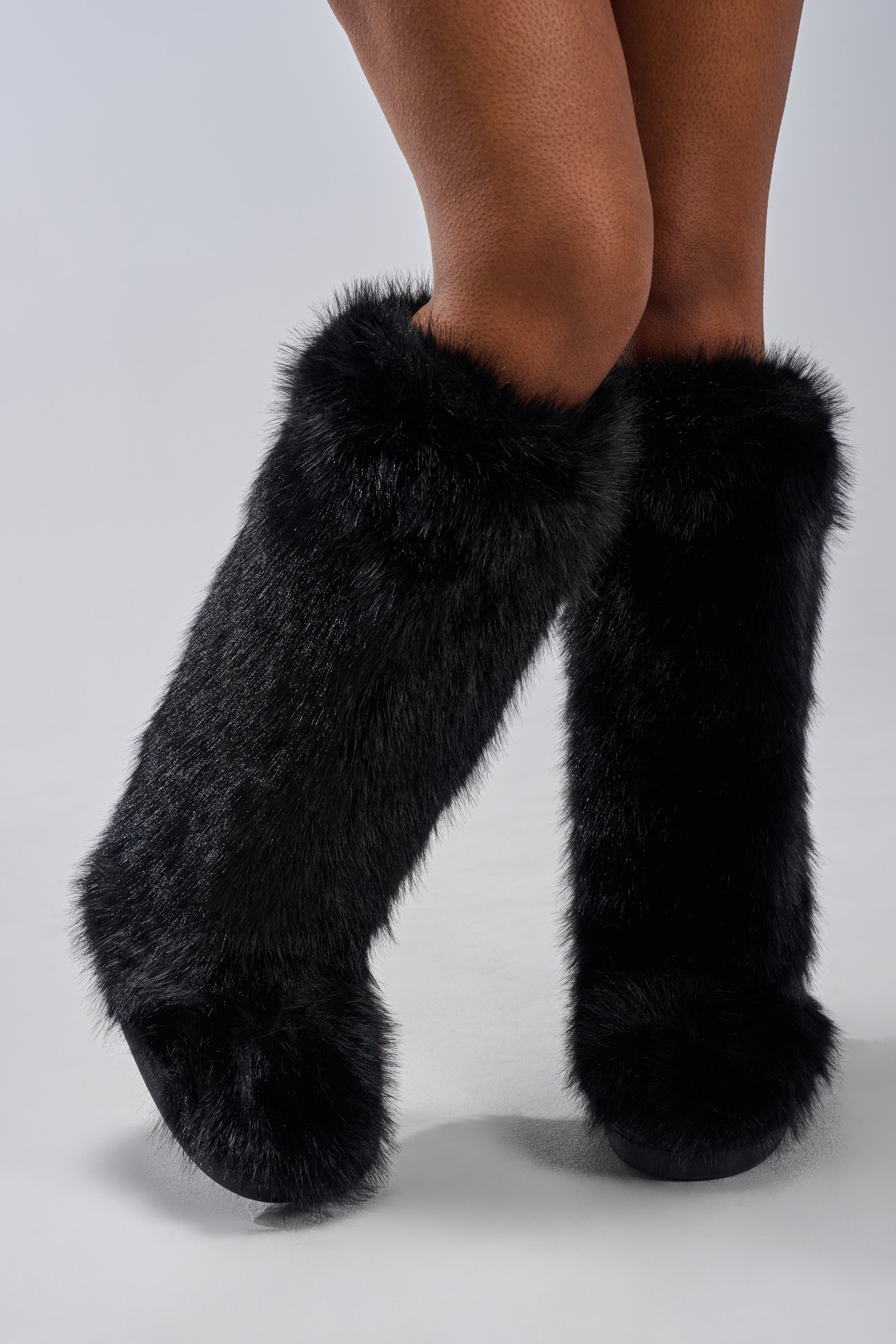 A person stands with legs crossed, wearing the AZALEA WANG EMIR BLACK FLAT FUR KNEE HIGH BOOT featuring tall, fluffy black faux fur and a flat sole. The boots cover the calves and the background is a plain light gray.