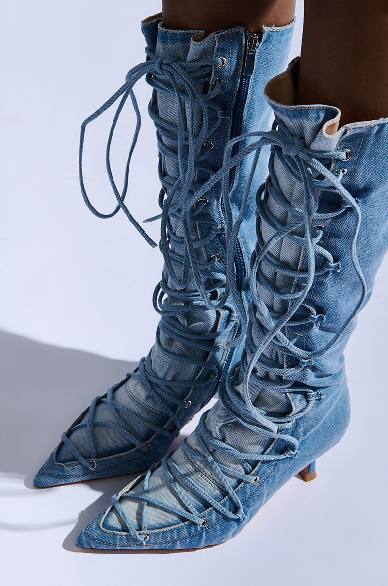 The AZALEA WANG EMMALINE DENIM STILETTO BOOT features knee-high shafts, pointed toes, and intricate lace-up detailing. The laces extend fully, creating an elaborate pattern. With small curved heels and a denim finish, they offer a stylish and casual look.