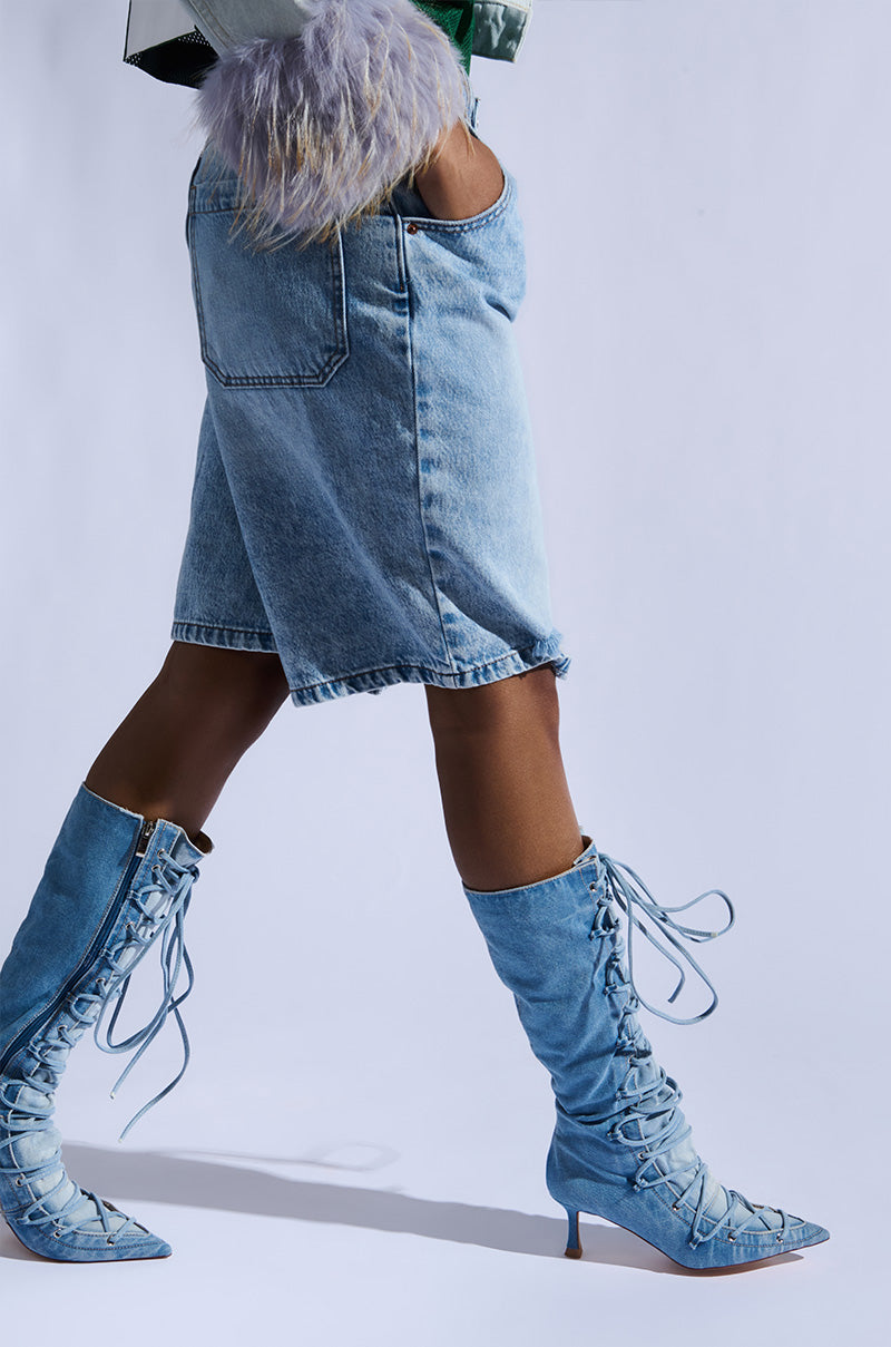 Striding confidently against a plain background in knee-high AZALEA WANG EMMALINE DENIM STILETTO BOOTS and a matching skirt, paired with a colorful top, the outfit exudes a casual yet trendy vibe that skillfully elevates classic denim with bold style choices.