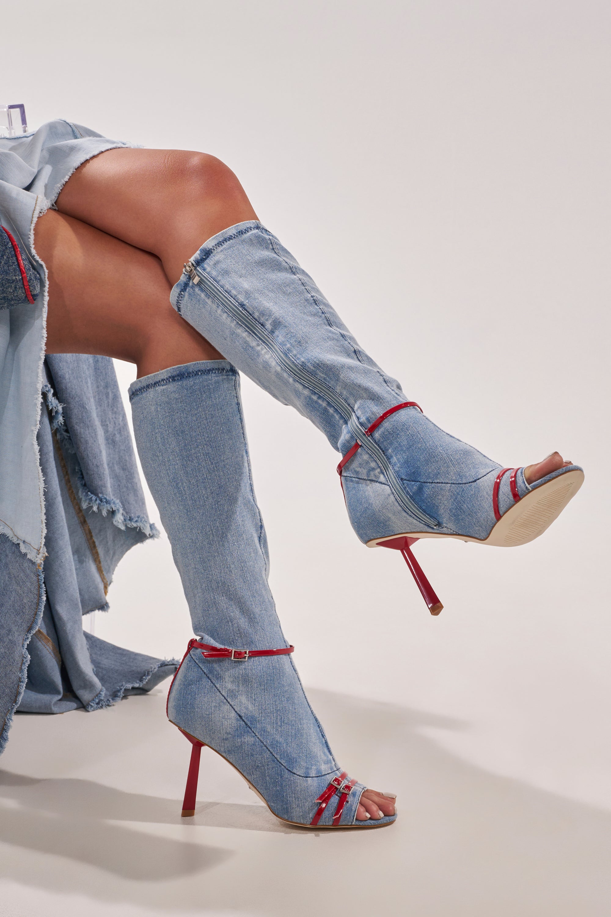 A person with crossed legs wears the AZALEA WANG EMIRA DENIM STILETTO OPEN TOE BOOT—knee-high denim boots with a red stiletto heel and peep toe—paired with a frayed-edge denim skirt or dress. 2