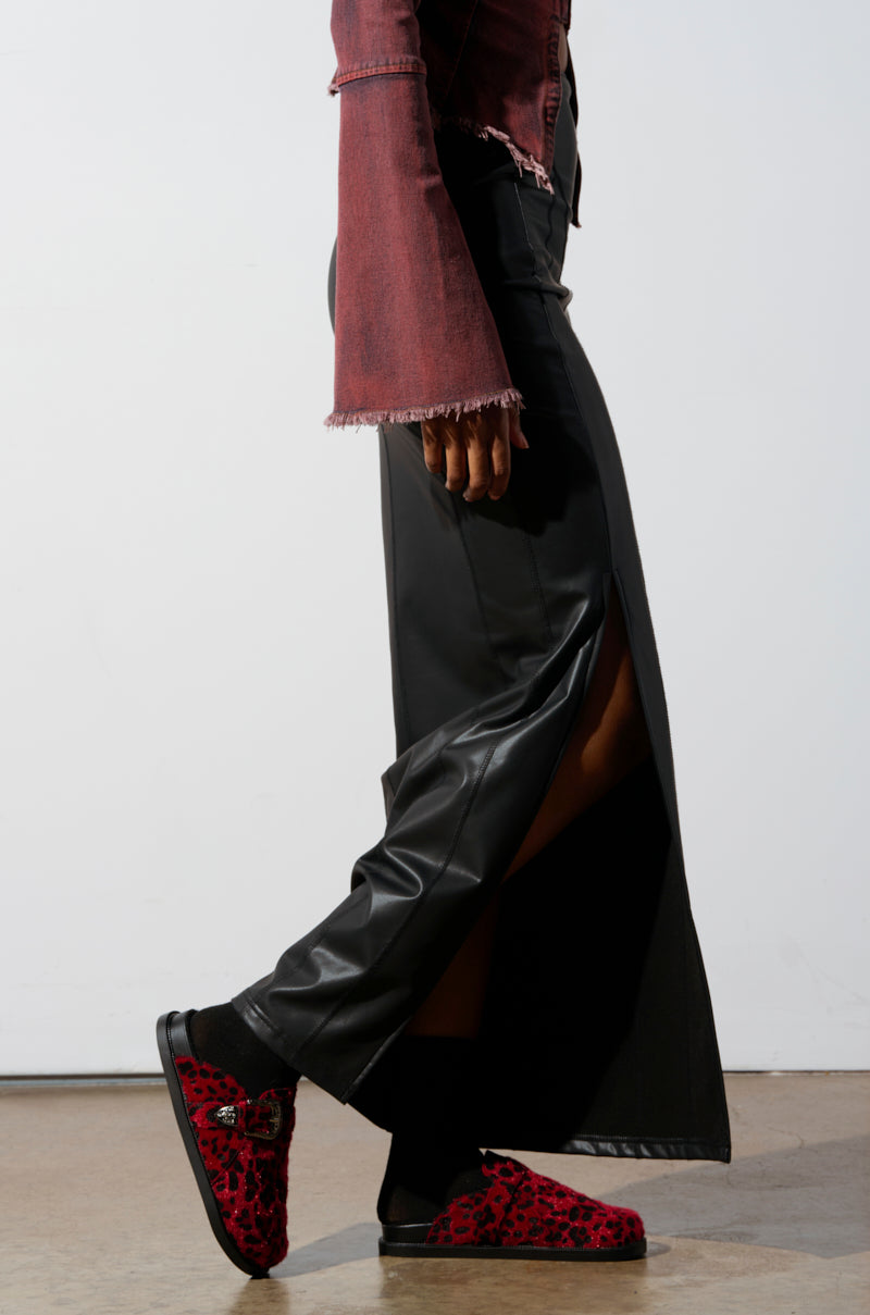 A person is dressed in a long black skirt featuring a high slit that reveals part of their leg, complemented by chic faux pony hair shoes in red leopard print. Their top is the AZALEA WANG ENDGAME RED LEOPARD jacket with frayed edges. They walk on a wooden floor set against a white background.