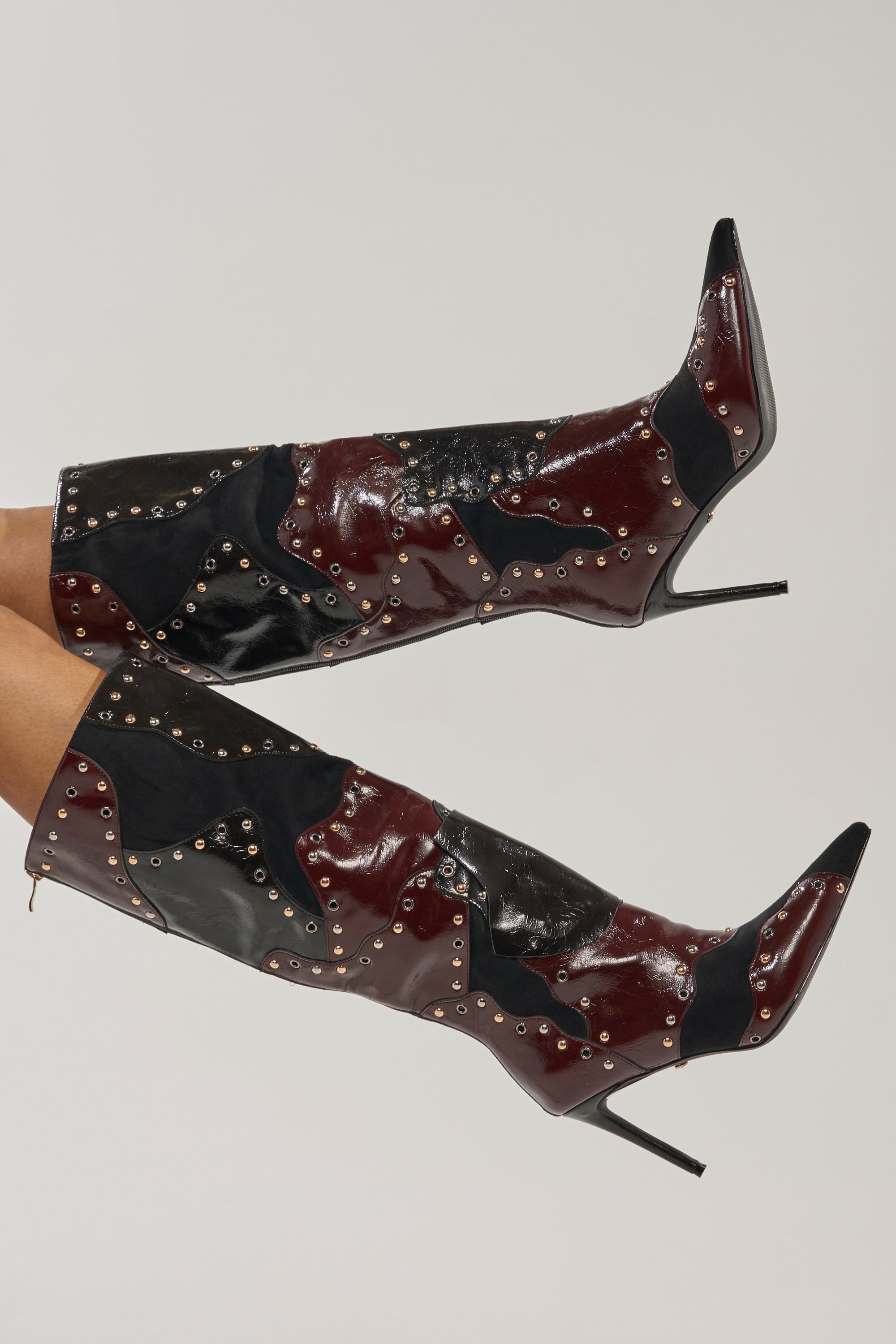 The AKIRA GOLD ENYAE MAHOGANY MULTI PATCHWORK STILETTO BOOT features a pointed toe, color block patchwork in black, burgundy, and dark blue faux leather, with metallic studs outlining each patch. 0