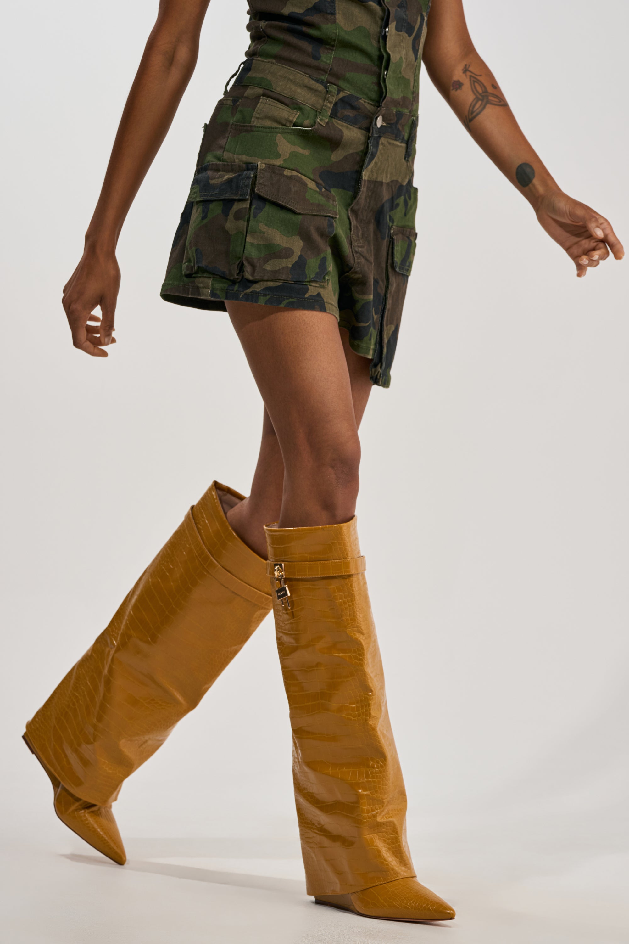 A person clad in a camo mini skirt and the AZALEA WANG ENZO MUSTARD CROC SHARK BOOT—a pair of oversized, foldover knee-high boots—walks, their lower half set against a plain backdrop for a striking statement streetwear moment.