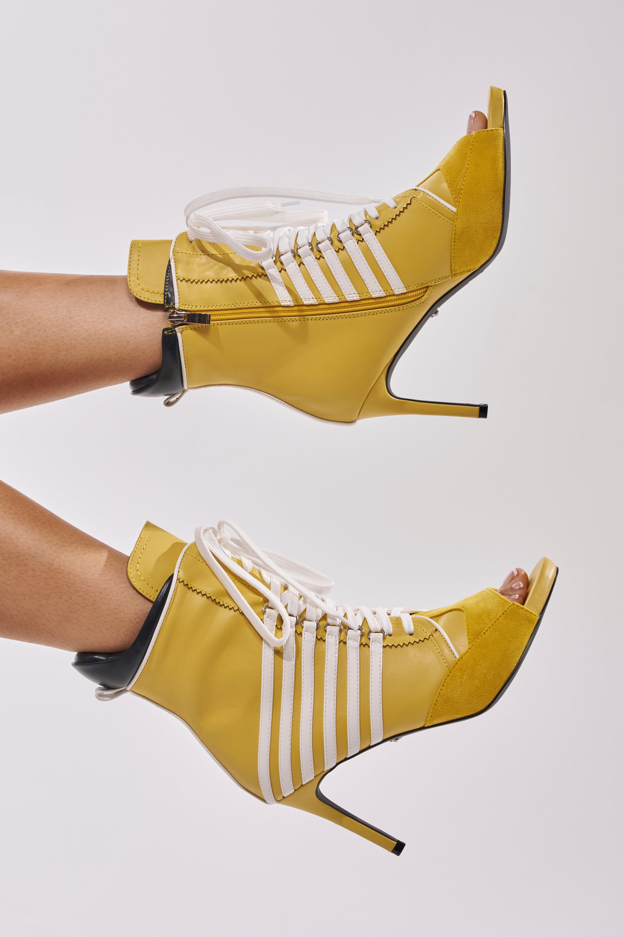 The AZALEA WANG EOWYN MUSTARD YELLOW OPEN TOE STILETTO BOOTIE features sporty side stripes, white laces, faux leather, black heel accents, and a pointed stiletto heel in bright yellow—worn with bare legs against a plain background.