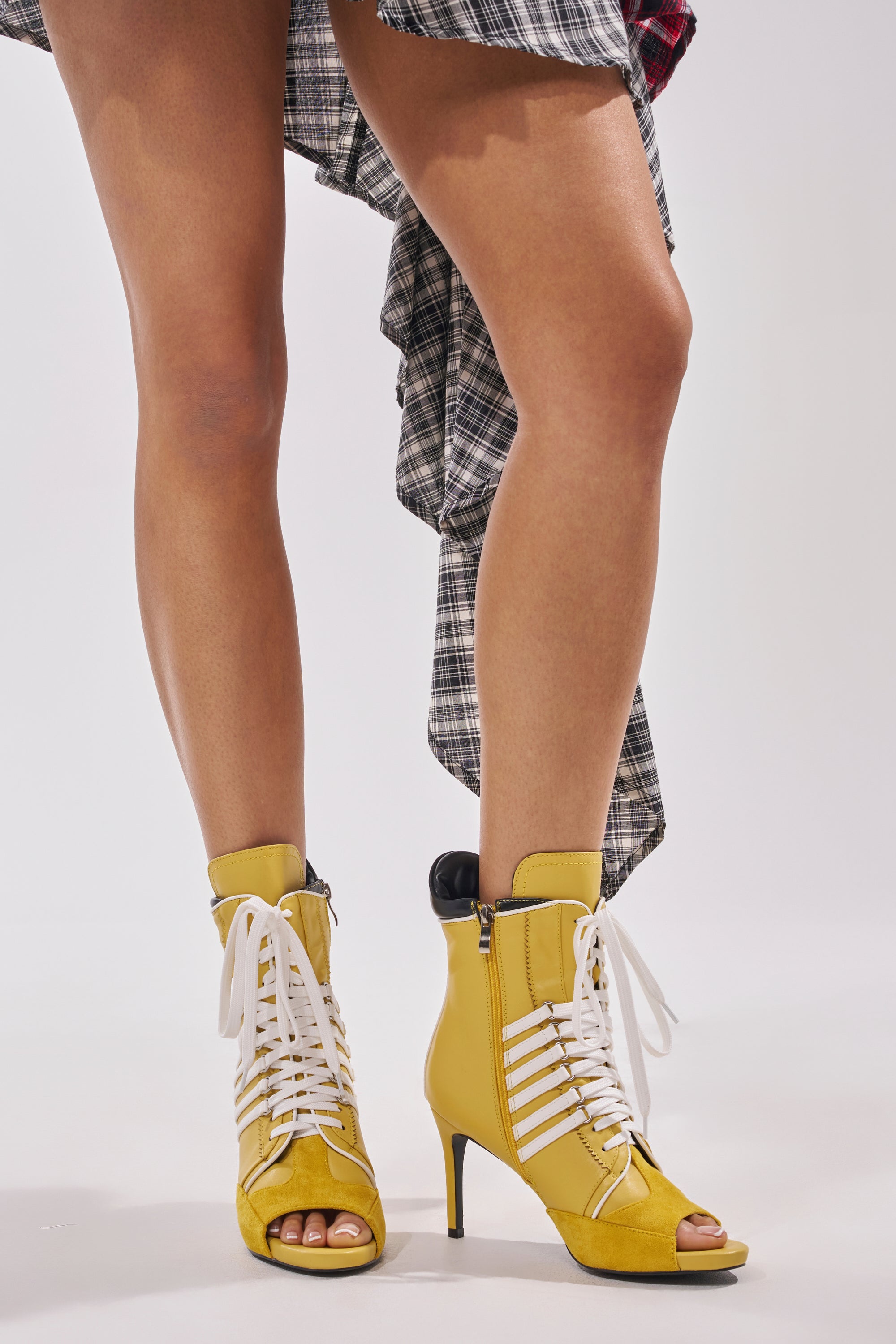 A person wearing a plaid skirt and the AZALEA WANG EOWYN MUSTARD YELLOW OPEN TOE STILETTO BOOTIE with white laces stands against a plain background, highlighting these striking, sporty booties. Only their legs and part of the skirt are visible.