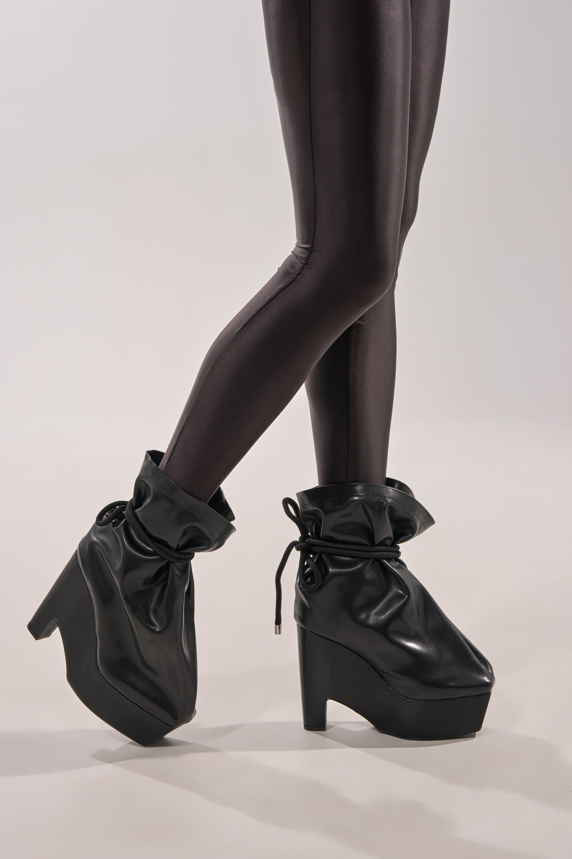 Wearing shiny black leggings and the AKIRA GOLD EPIGRAM PU SLOUCH CHUNKY HEEL BOOTIE with loose, slouchy tops featuring drawstring details, a person stands on a light, neutral background. 1