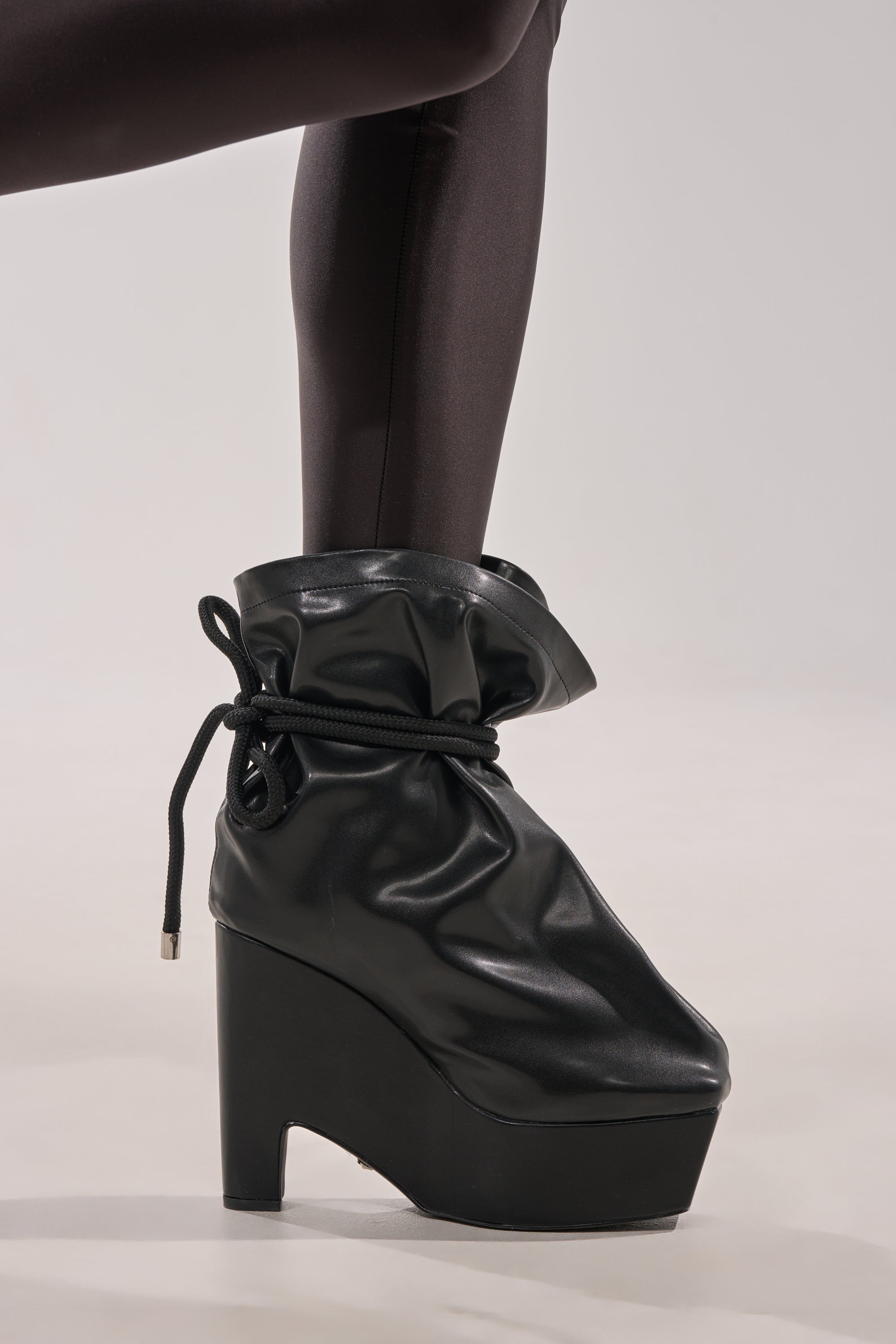 A person wears shiny black leggings and AKIRA GOLD EPIGRAM PU SLOUCH CHUNKY HEEL BOOTIE—high platform booties made of glossy faux leather with a drawstring detail around the ankle. 2