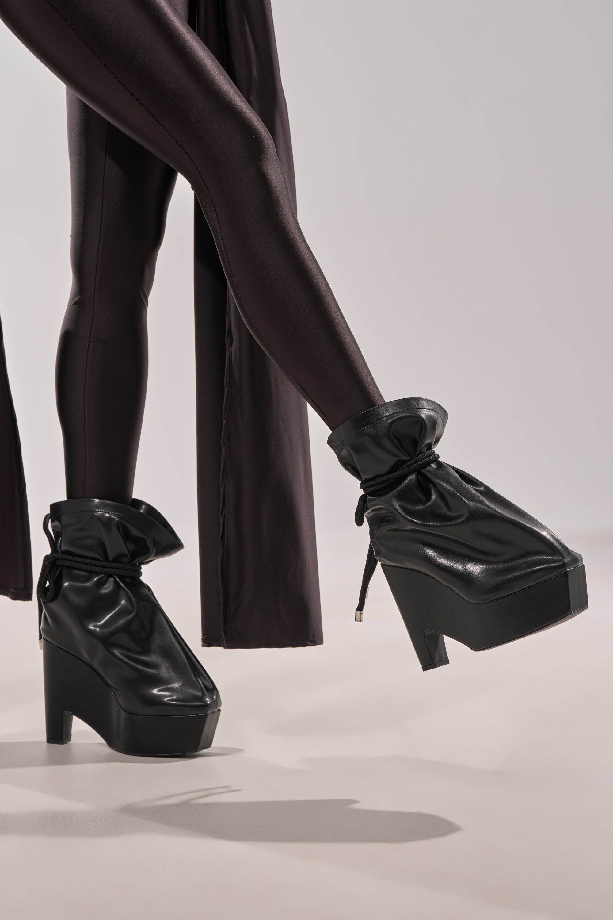 A person wearing shiny black leggings and the AKIRA GOLD EPIGRAM PU SLOUCH CHUNKY HEEL BOOTIE, featuring a unique ruched, baggy faux leather design, stands against a plain light gray background. 7