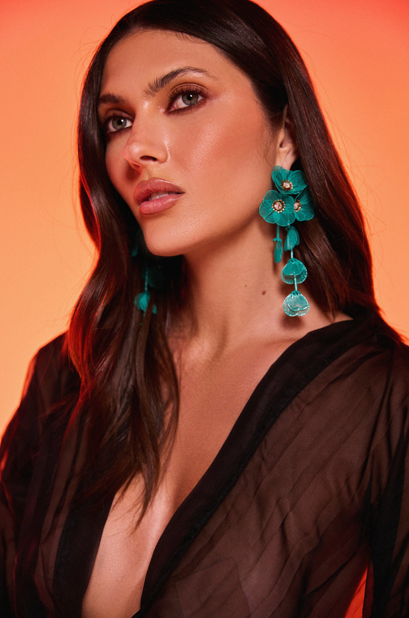 A woman with long dark hair models the TOTEALLY EARRING—bold, botanical statement earrings—paired with a sheer black top, posing confidently against a warm orange background. 1