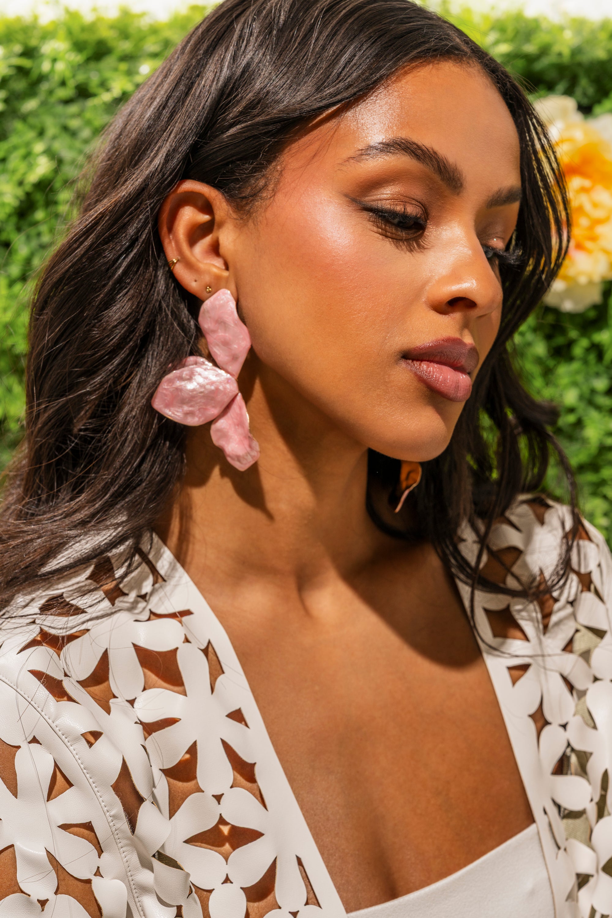 A woman with long dark hair poses outdoors among green foliage and yellow blooms, wearing a white floral cutout top and the PRETTY IN PINK EARRING—lightweight enamel earrings shaped like large pink flowers, perfect for vacation style. 0