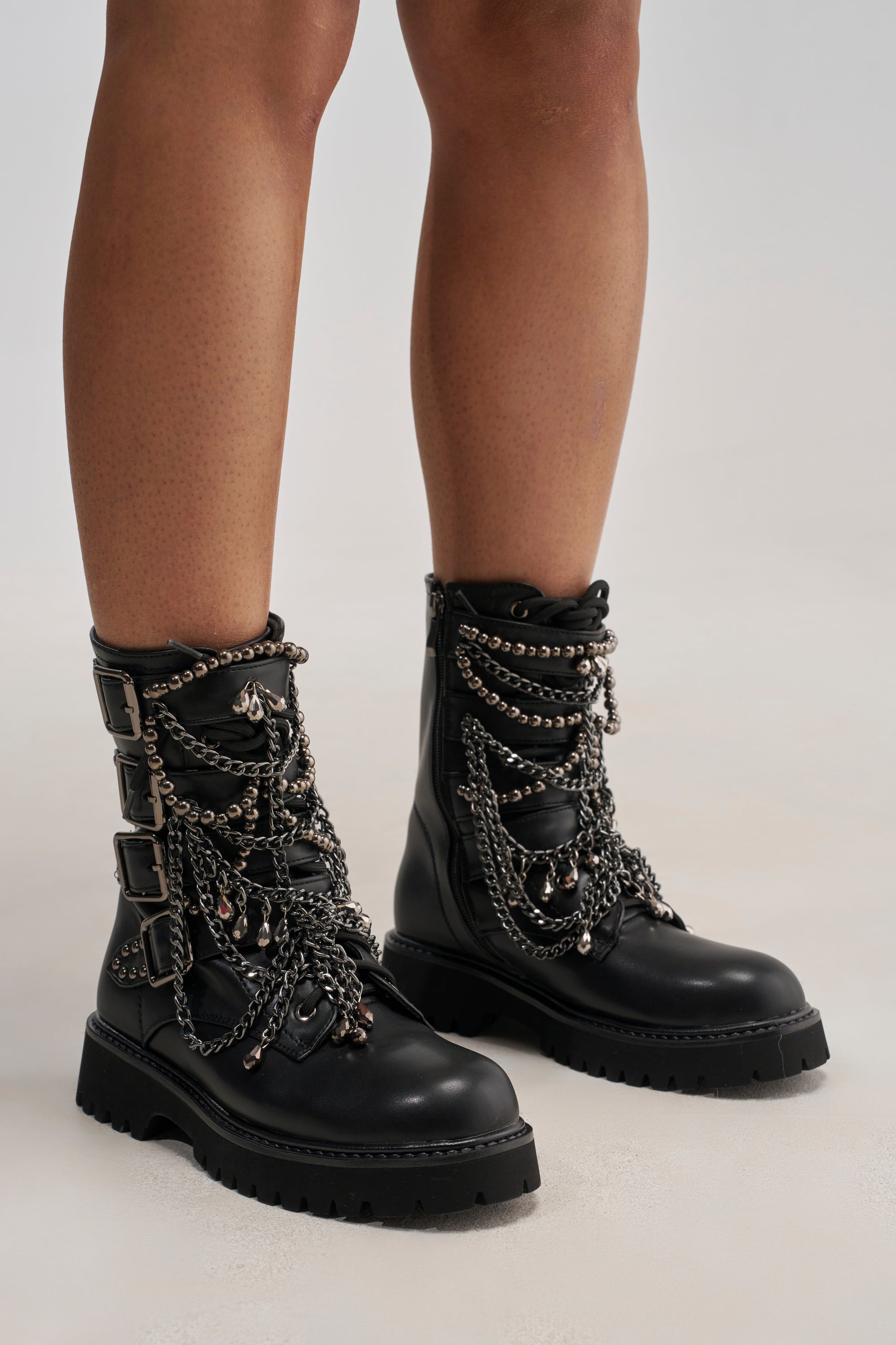 A person stands on a light-colored floor, wearing the AZALEA WANG ERIN EMBELLISHED COMBAT BOOTIE in black faux leather, adorned with multiple silver chains and buckles. Only the lower legs and boots are visible.